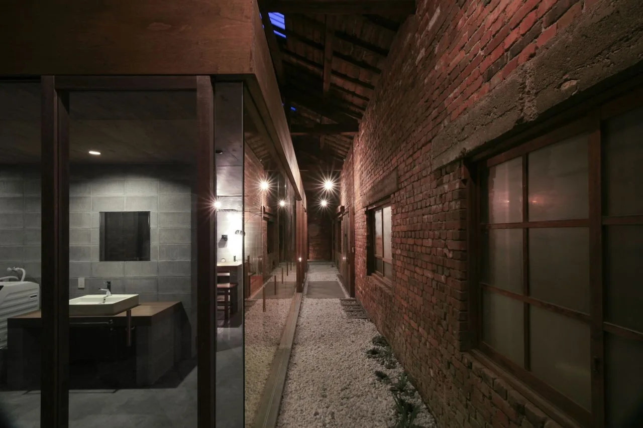 An indoor corridor features an exposed brick wall, gravel flooring, and a glass-enclosed bathroom with a concrete block wall and wooden vanity.