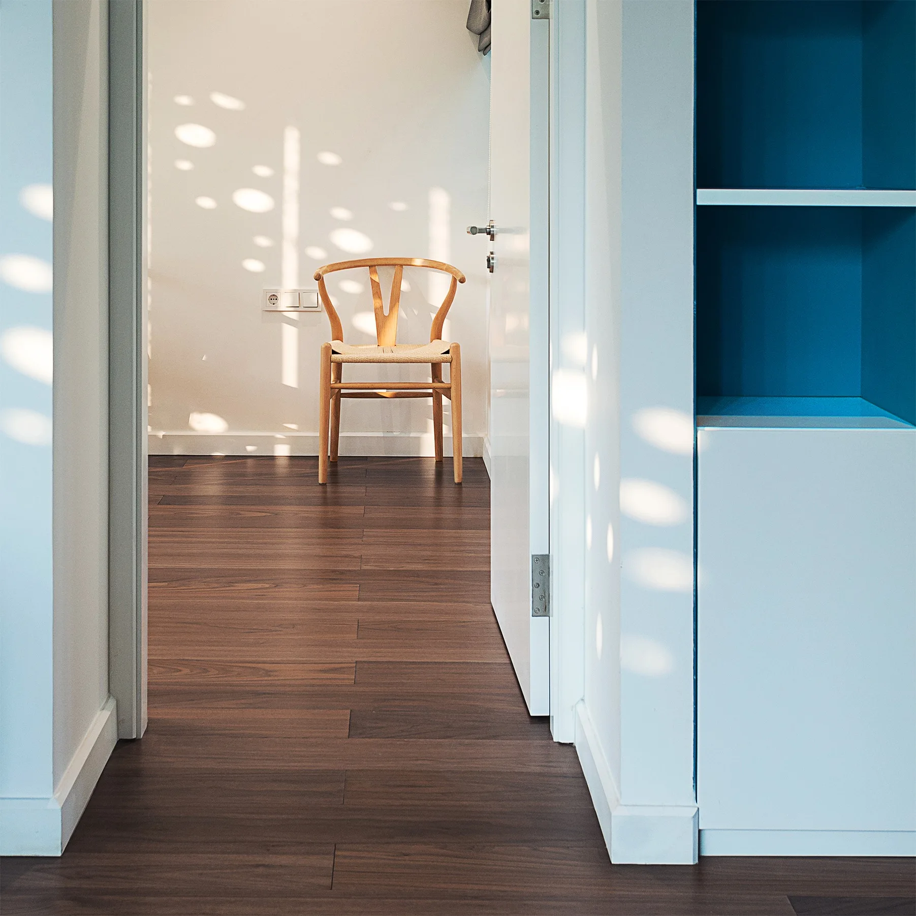 A view through a white-walled hallway with dark wood floors, featuring a classic wooden wishbone chair under dappled sunlight and a blue built-in shelf.