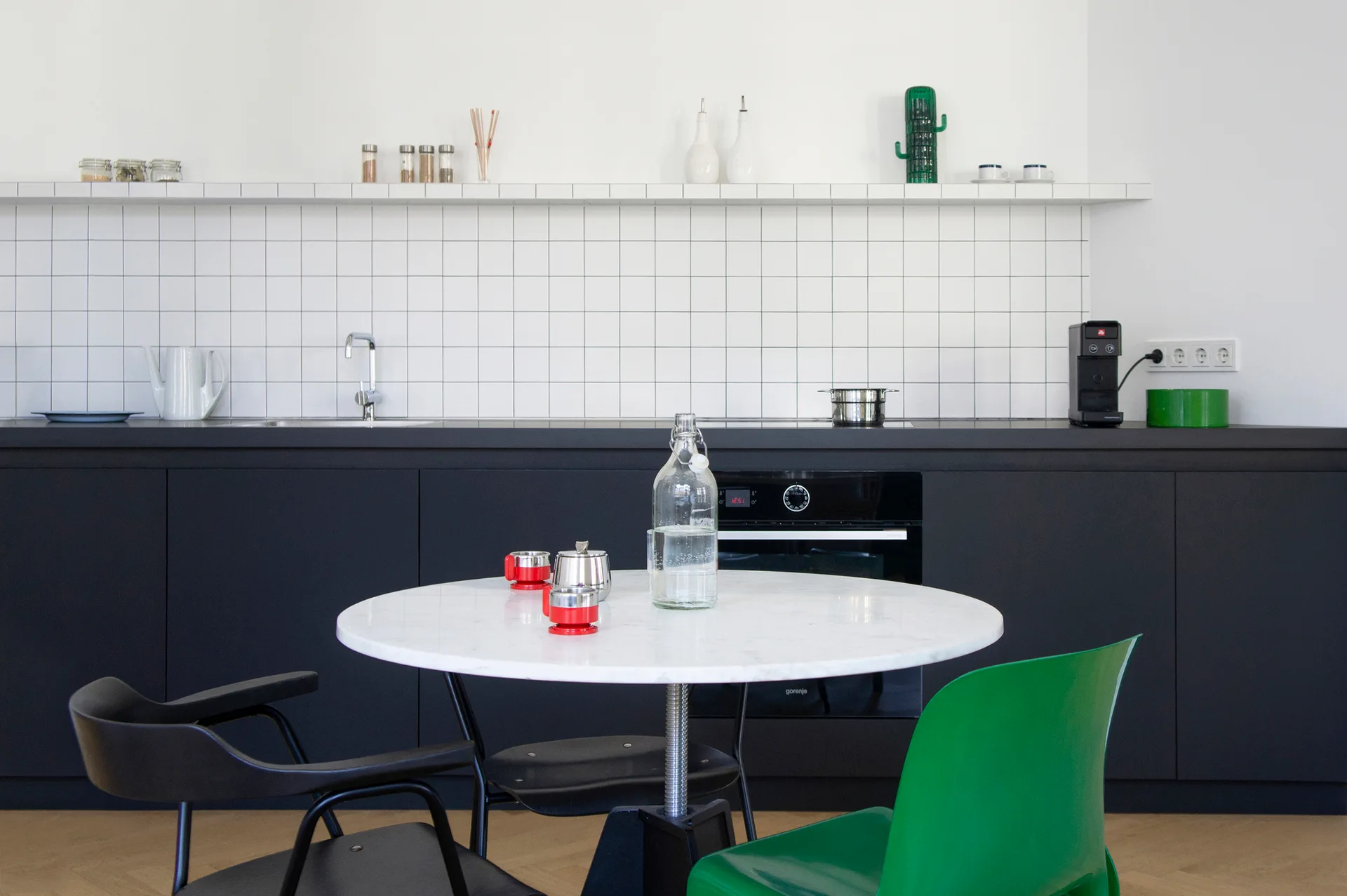 A modern kitchen featuring matte black cabinets, a white square tile backsplash, and a round marble dining table with mismatched black and green chairs.