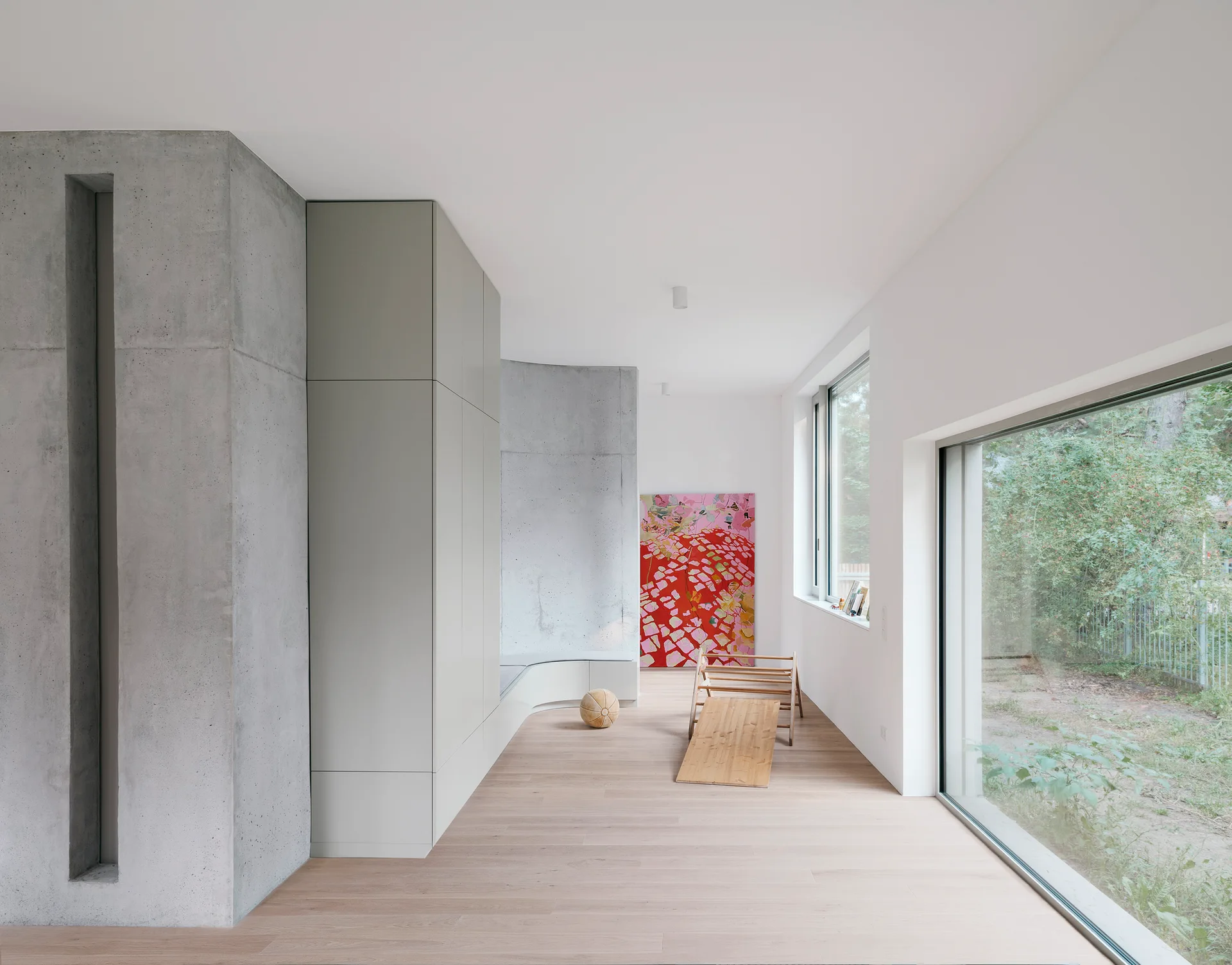 A minimalist hallway features light wood floors, a raw concrete pillar, sleek gray storage cabinets, and large windows looking out to trees.