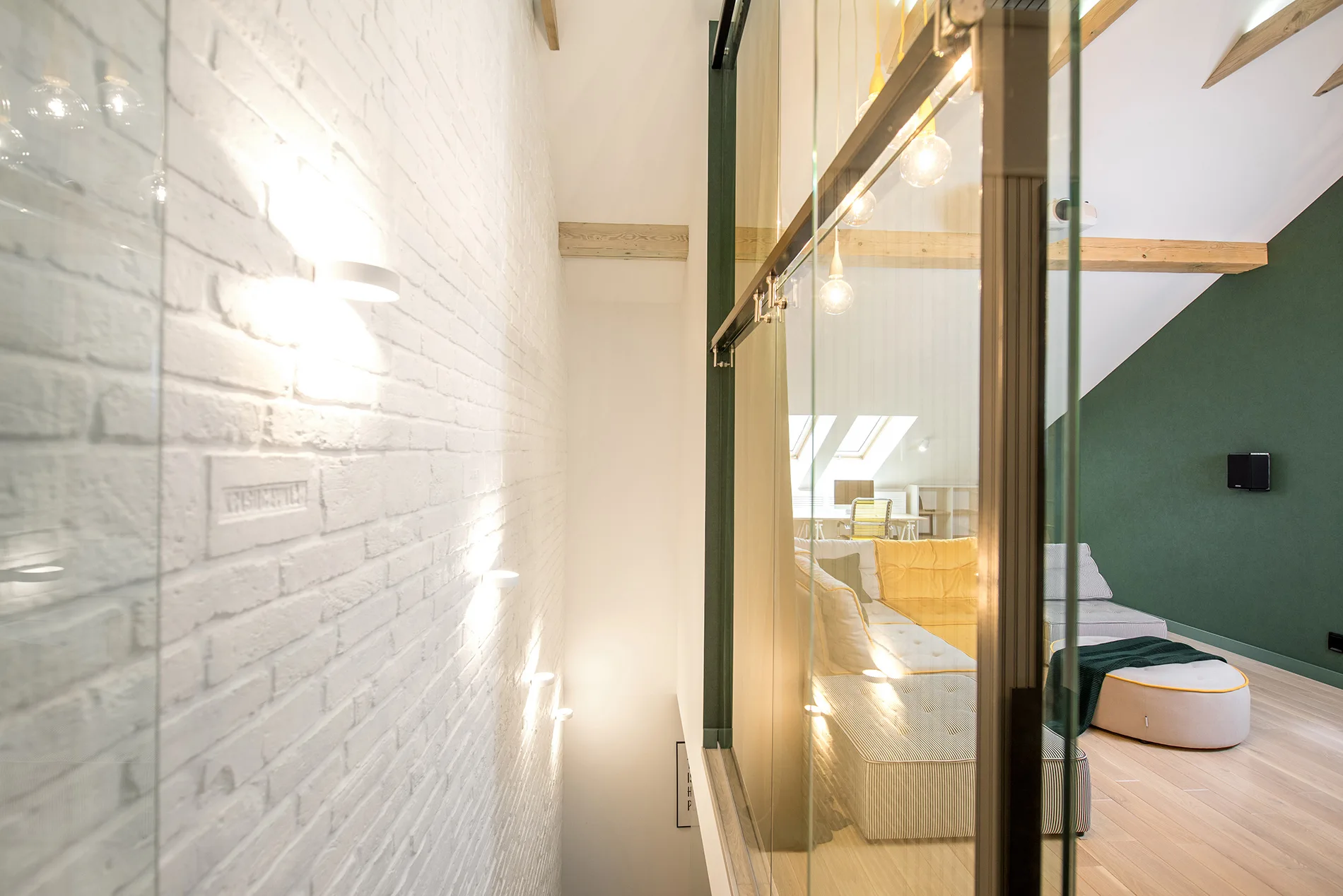 A view through a glass sliding partition reveals a loft living room with a white brick wall, exposed ceiling beams, and a yellow modular sofa.