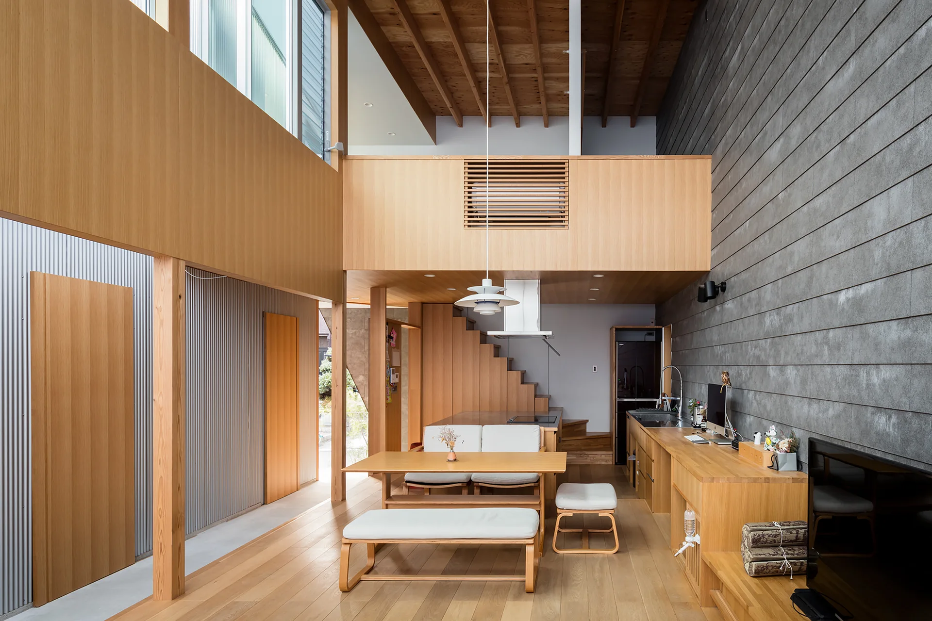 A modern minimalist dining area with light wood furniture, a grey plank wall, and double-height ceiling with exposed rafters.