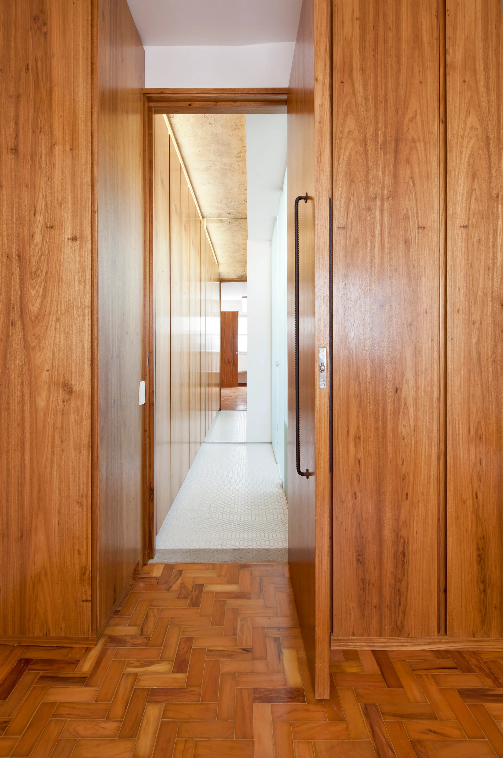 A narrow hallway featuring warm wood built-in cabinetry, herringbone parquet flooring, and a transition to white mosaic tile under a lit ceiling.