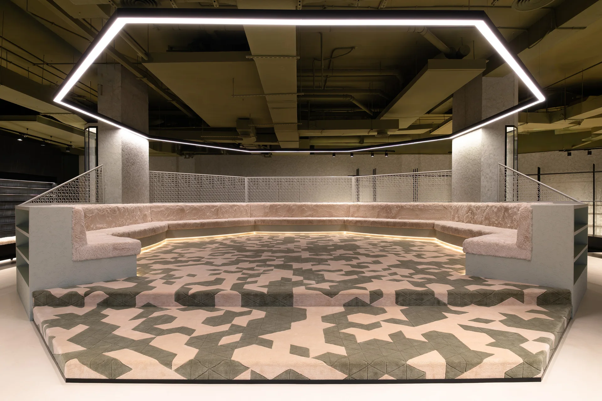 A hexagonal raised platform features fuzzy built-in bench seating, a large geometric pattern carpet, and a matching suspended light fixture overhead.