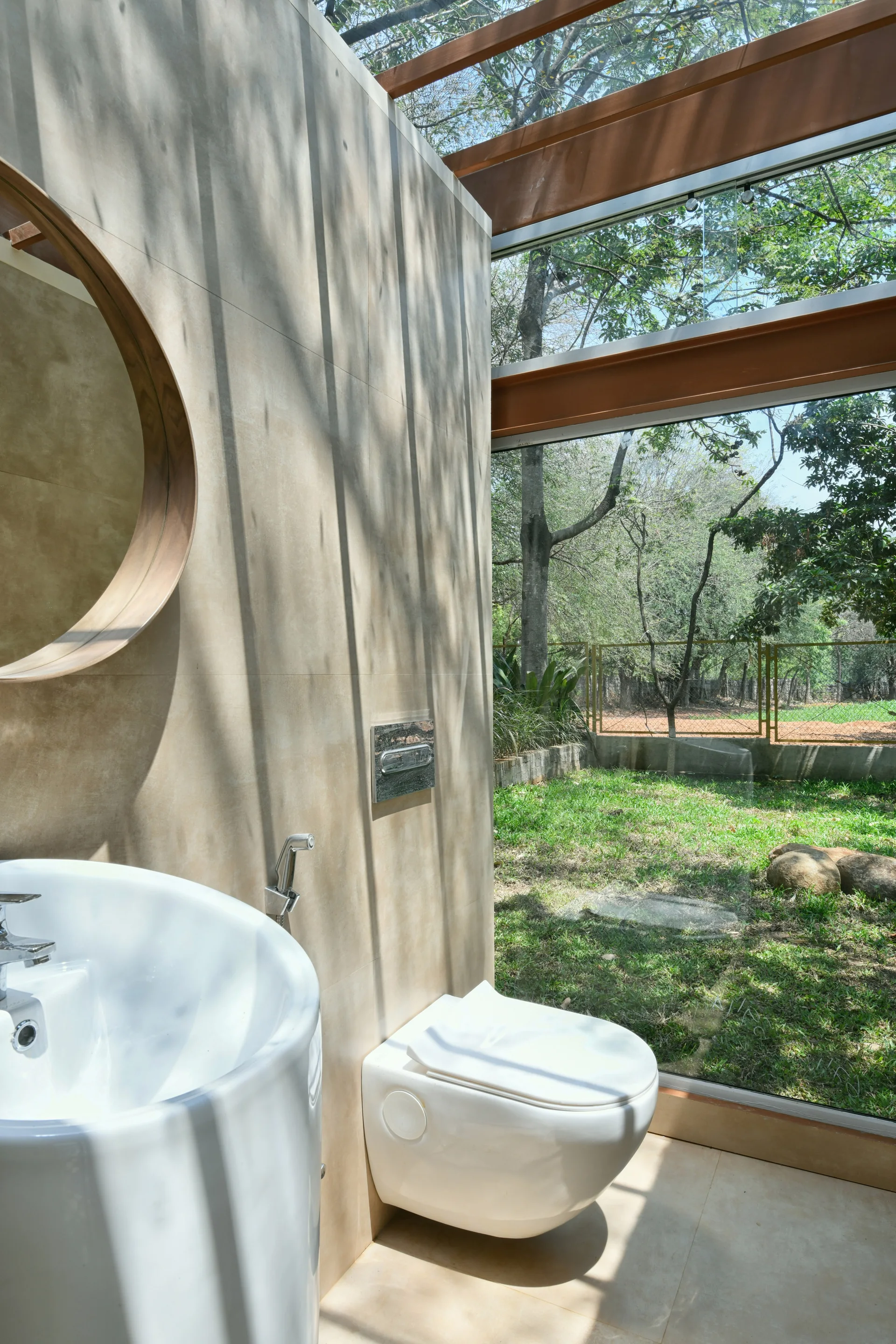 A minimalist bathroom features a wall-mounted toilet and sink next to a floor-to-ceiling glass window showcasing a green garden.