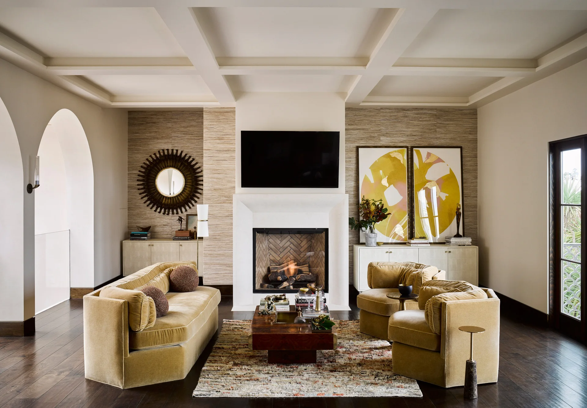 A symmetrically arranged living room featuring a central stone fireplace, golden velvet seating, a coffered ceiling, and textured wall panels.