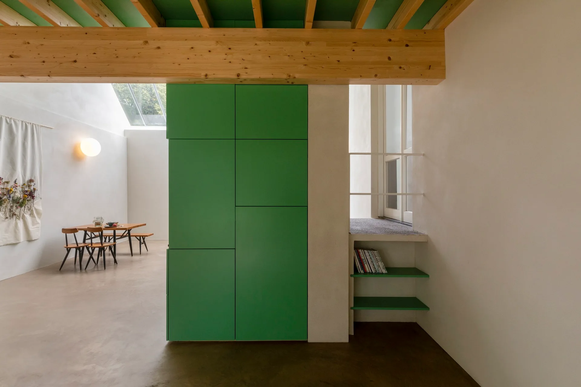 A large green storage cabinet stands below exposed wood ceiling beams in a minimalist room with white plaster walls and grey concrete floors.