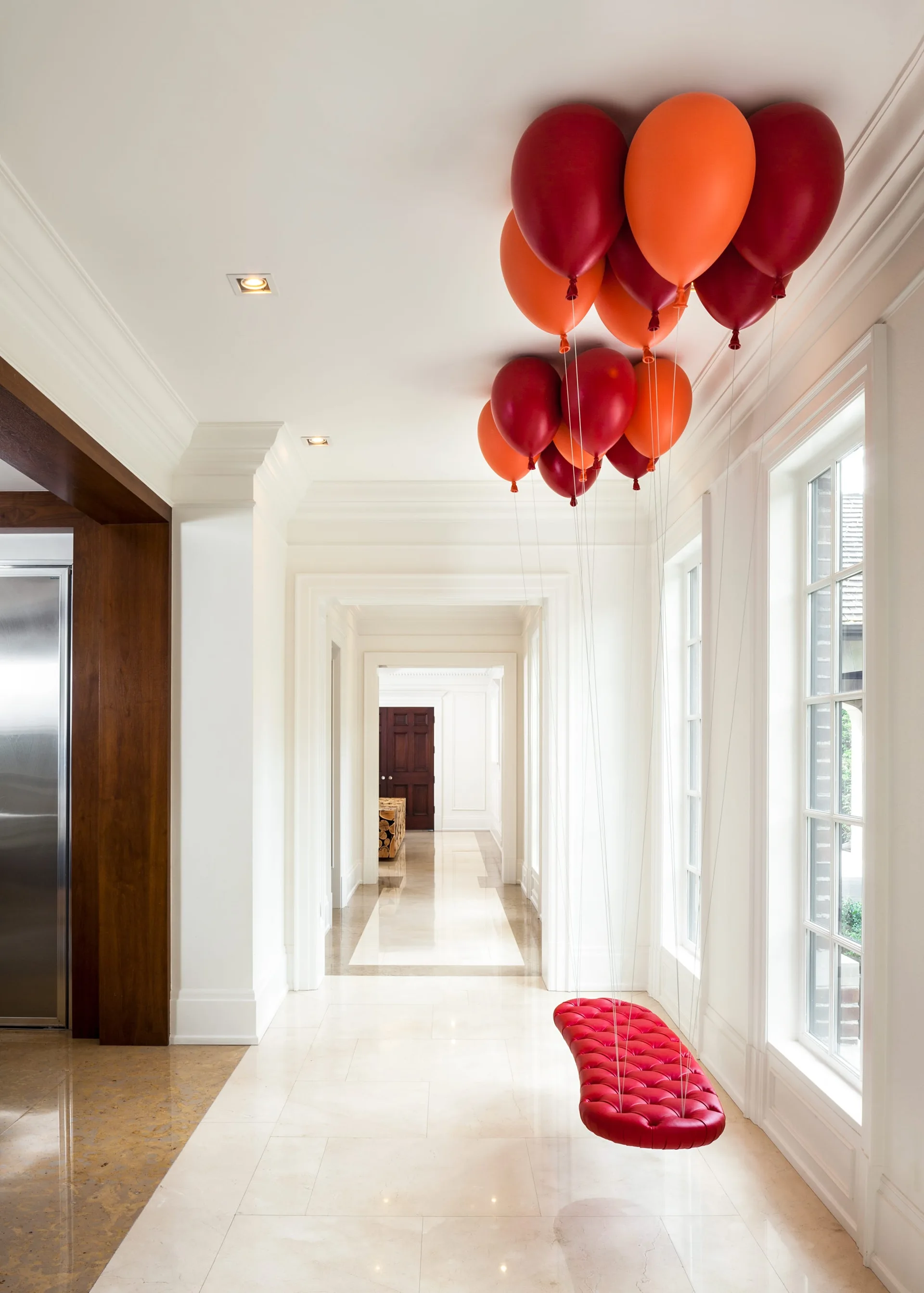 A long white hallway with a polished floor features a tufted red bench suspended from a cluster of red and orange balloons.