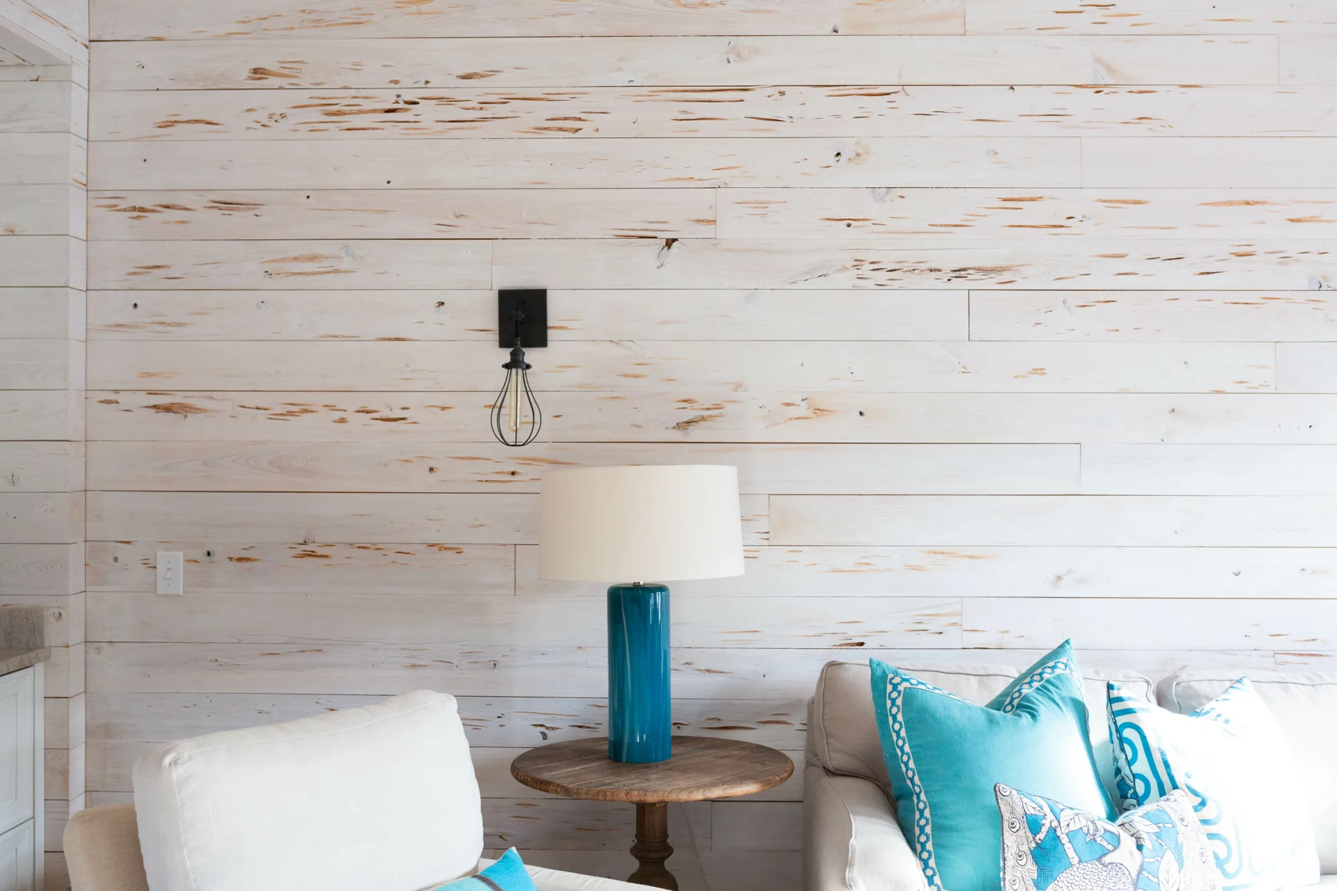 A living room corner features a white horizontal shiplap wall, a round wood side table with a blue ceramic lamp, and a black cage sconce.