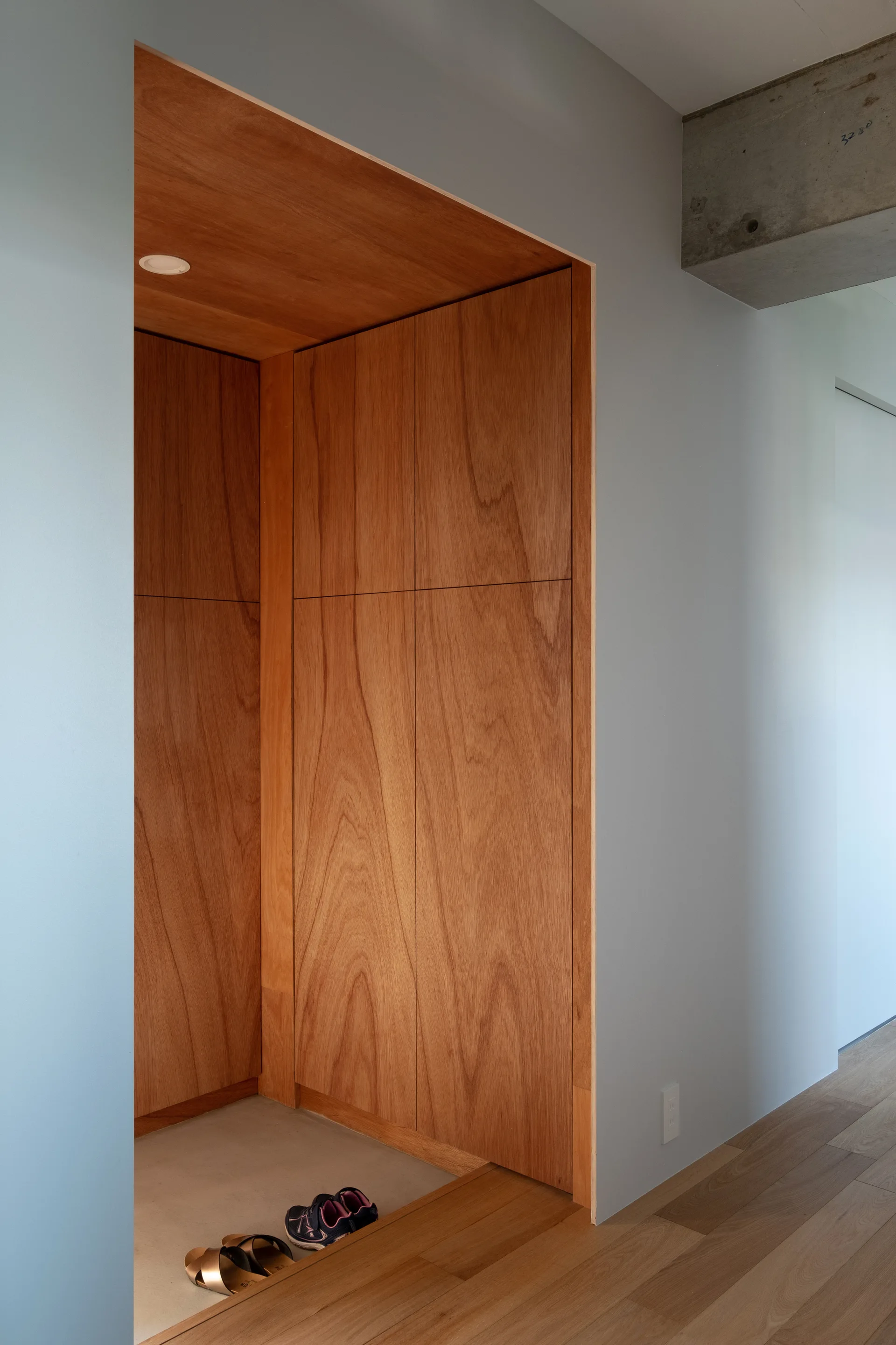 A recessed entryway featuring wood storage cabinets, light grey walls, a lower concrete floor area with shoes, and light wood plank flooring.