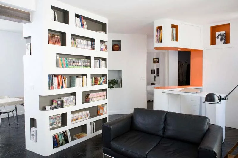 A modern living room features a large white room divider with asymmetrical recessed bookshelves next to a black leather sofa and dark floors.