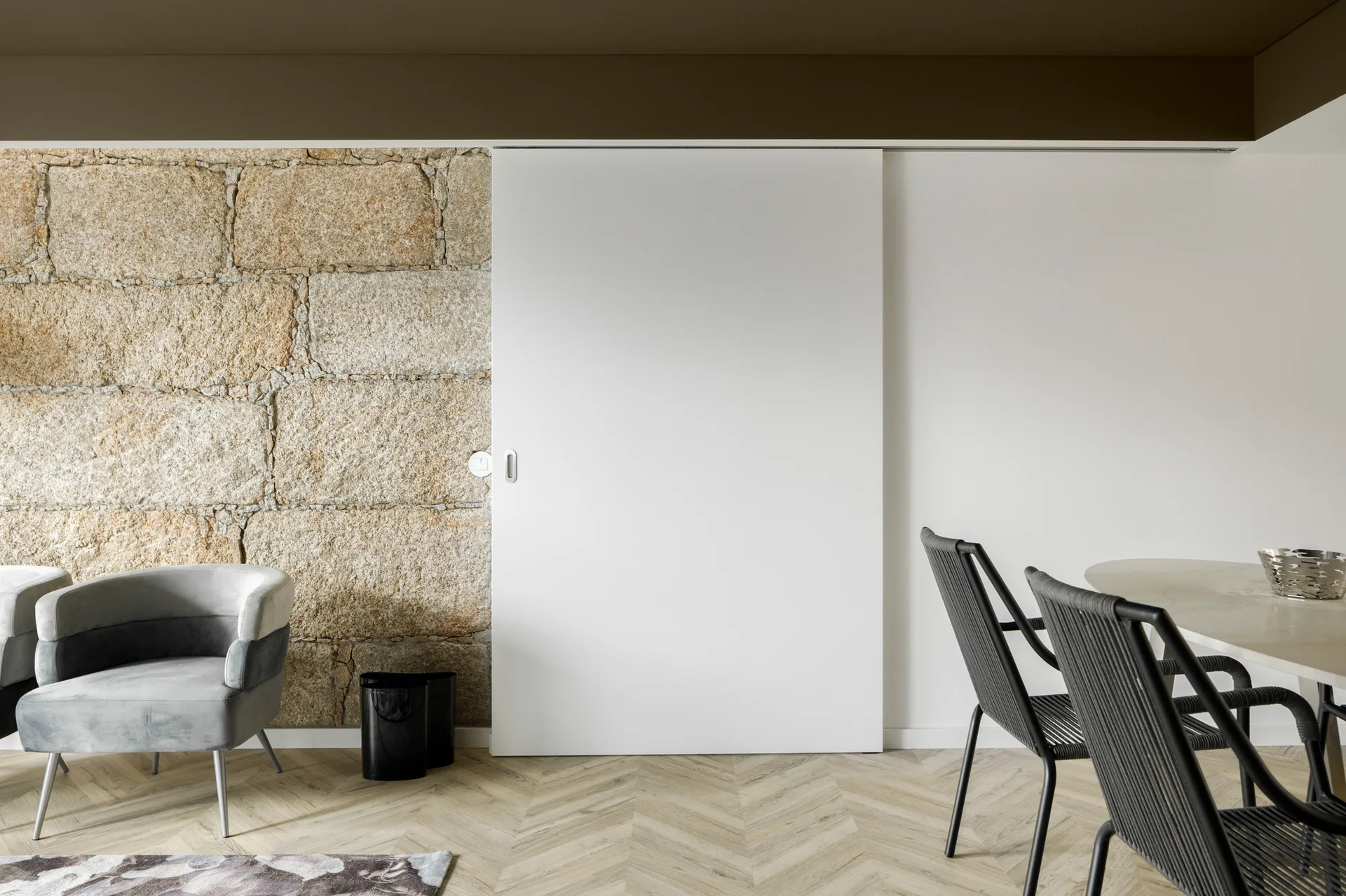 A dining area featuring a rustic stone wall next to a large white sliding door, with black woven chairs, a grey armchair, and herringbone wood floors.