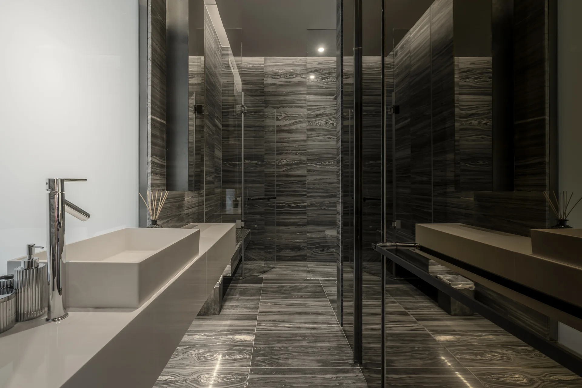 A modern bathroom featuring dark veined stone wall and floor tiles, a floating white vanity with a chrome faucet, and a glass walk-in shower.