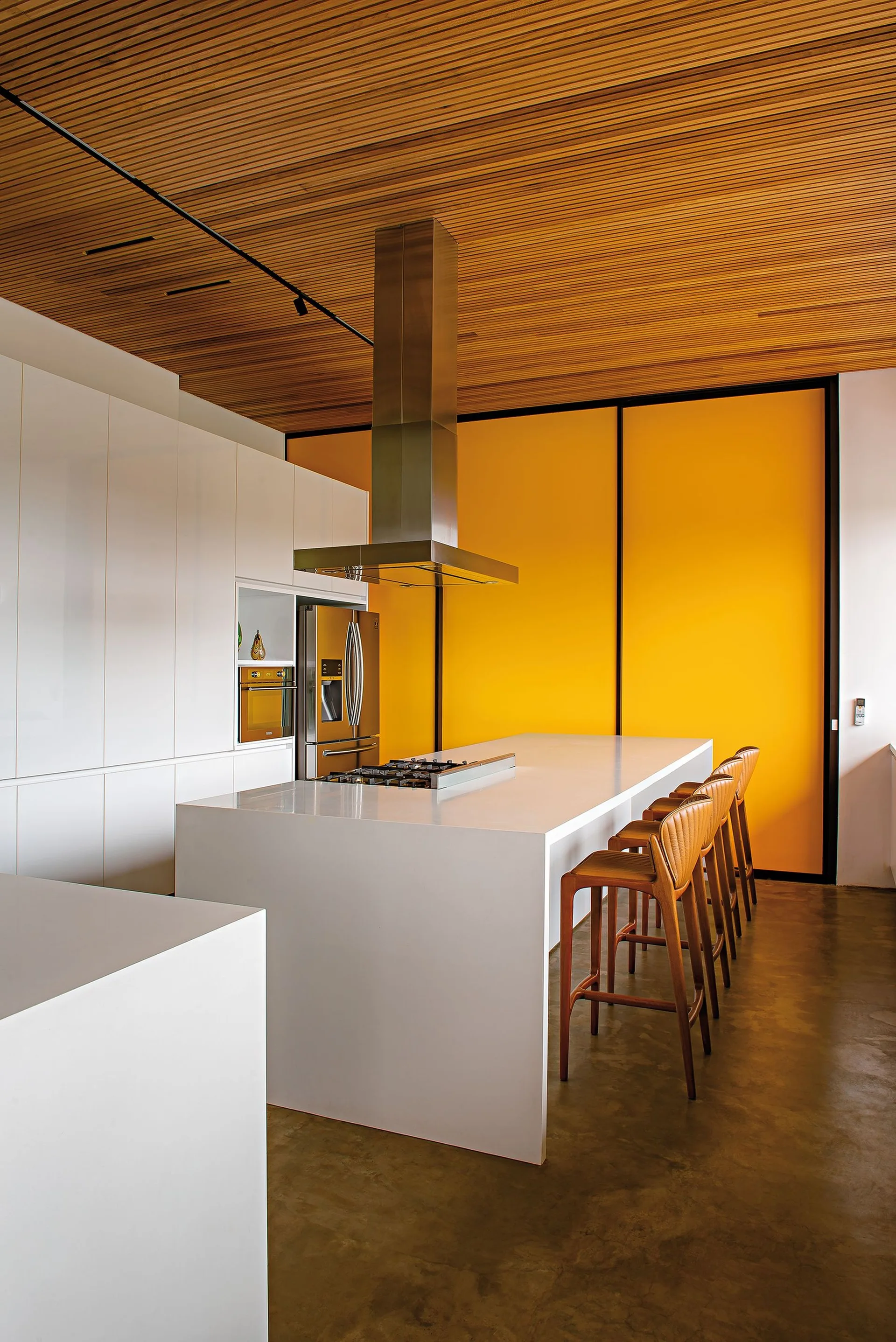 A minimalist white kitchen features a large island with wooden stools under a slatted wood ceiling next to a vibrant yellow sliding accent wall.
