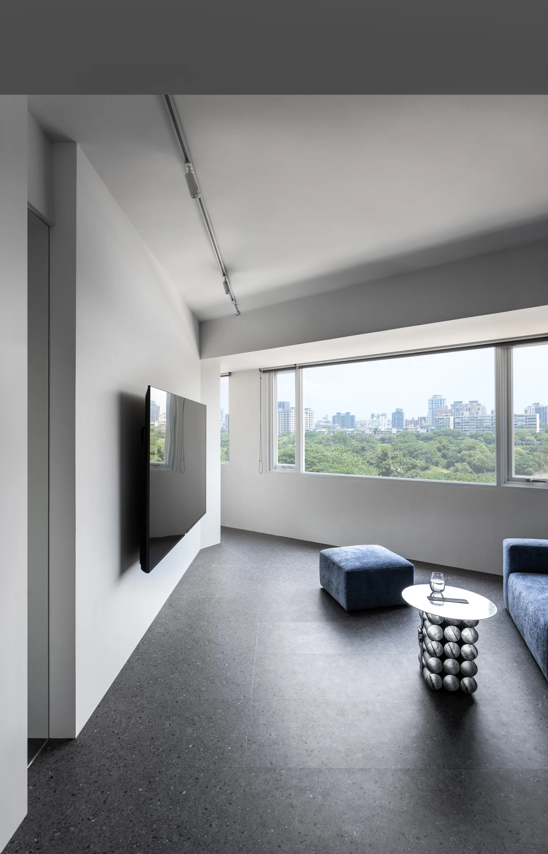 A minimalist living room features dark speckled tile flooring, a blue sofa and ottoman, a silver sculptural side table, and a wide window.