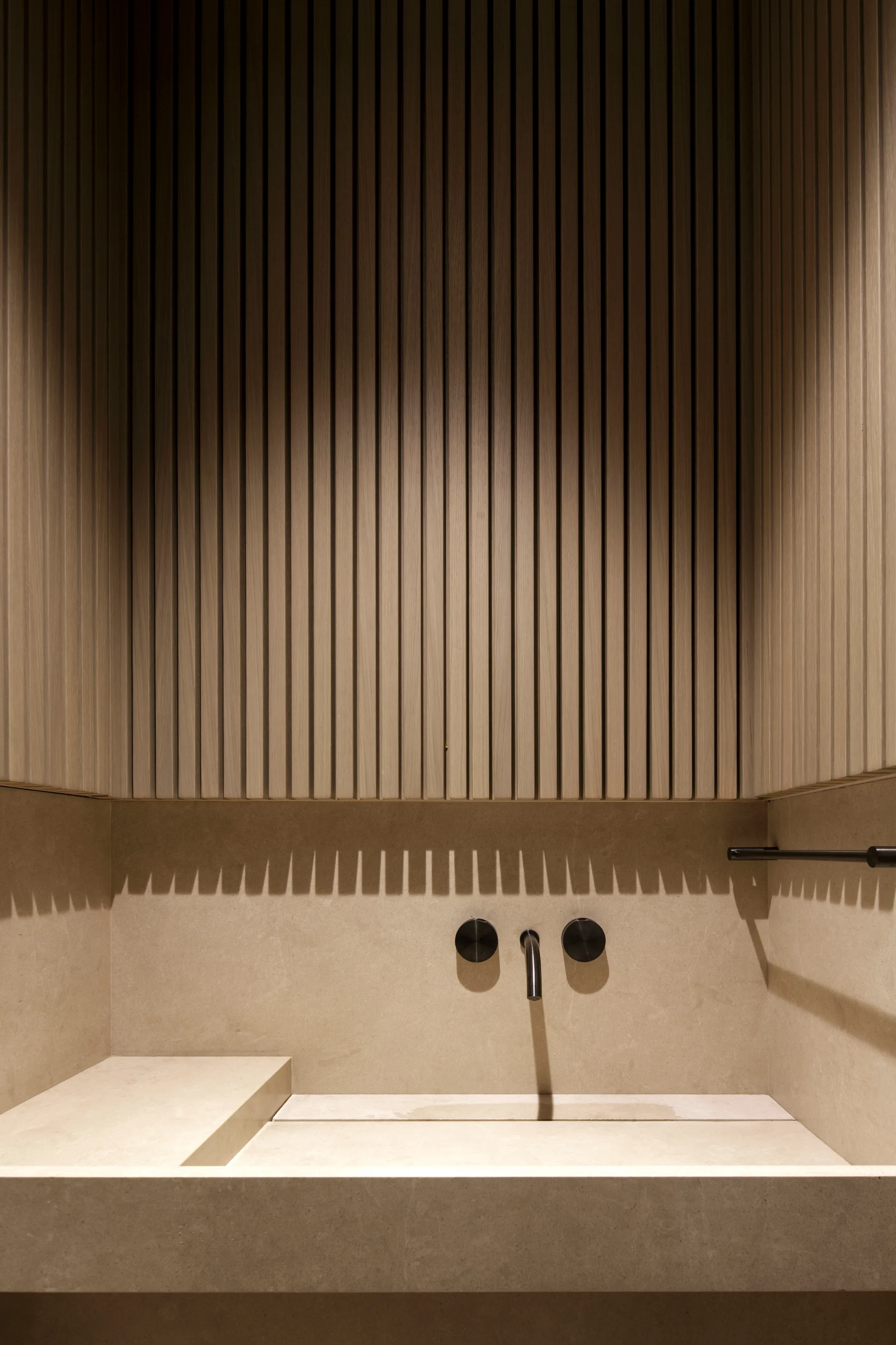 A beige stone bathroom vanity with an integrated sink and wall-mounted black faucet sits beneath vertical slatted wood wall paneling.