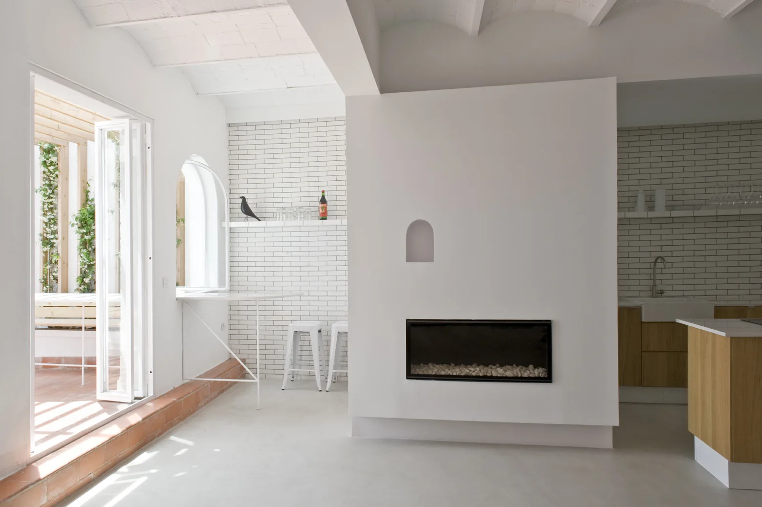 A minimalist white room with a central fireplace partition wall, an open arched window, and kitchen cabinetry in the background.