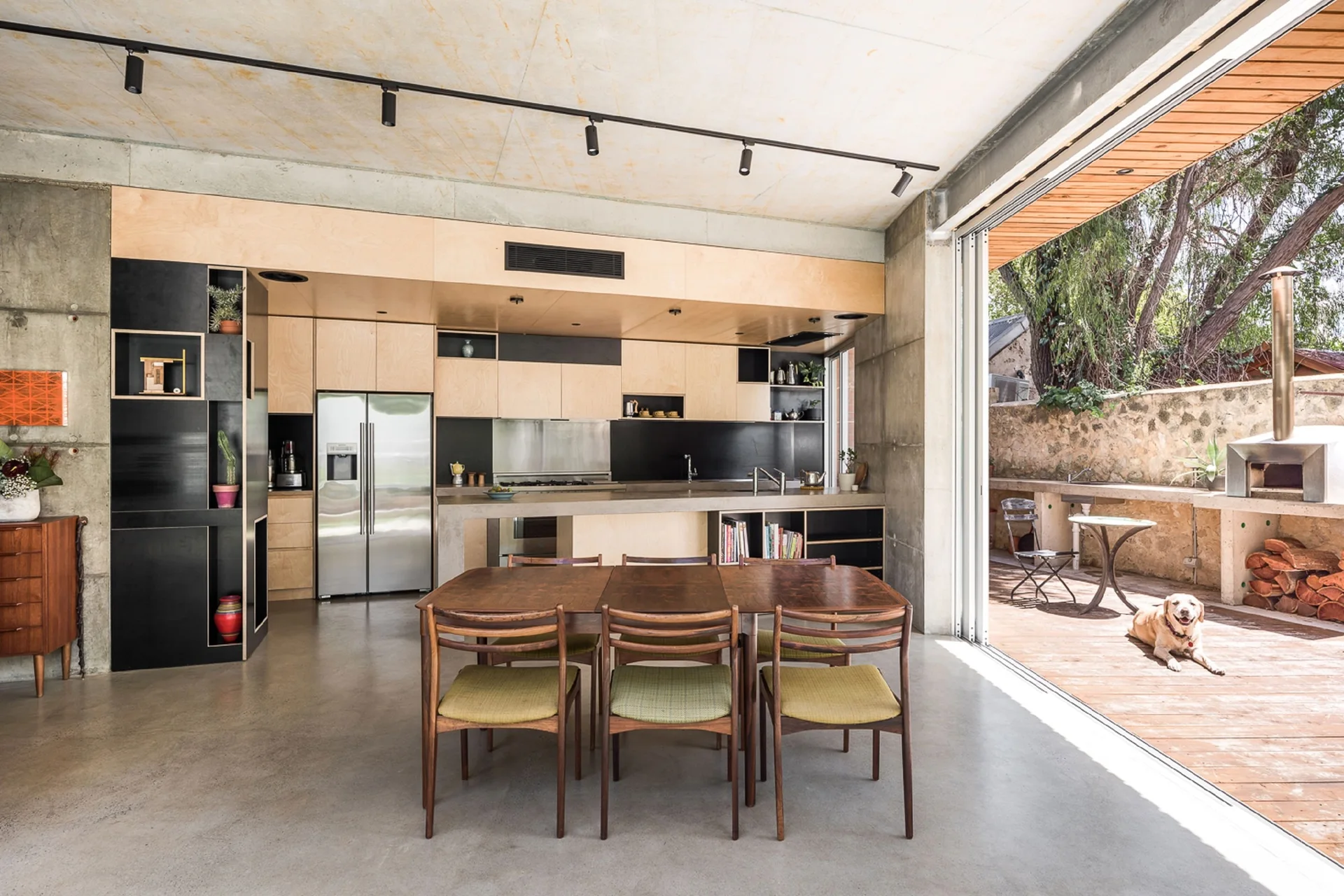 A modern industrial kitchen features plywood cabinets, polished concrete floors, and a wooden dining table next to a sliding glass door opening to a deck.