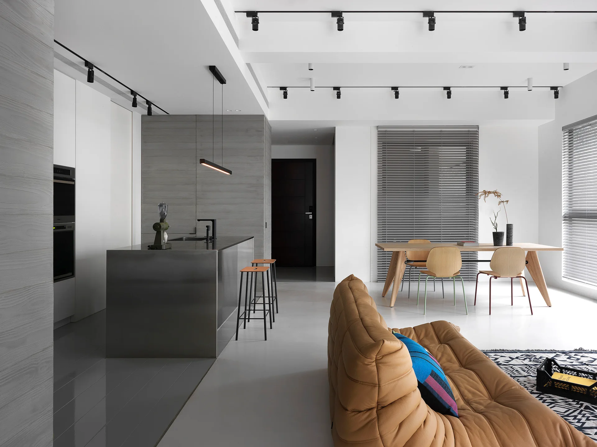 A modern open-plan interior featuring a metallic kitchen island, a tan modular leather sofa, a wooden dining table, and black ceiling track lights.