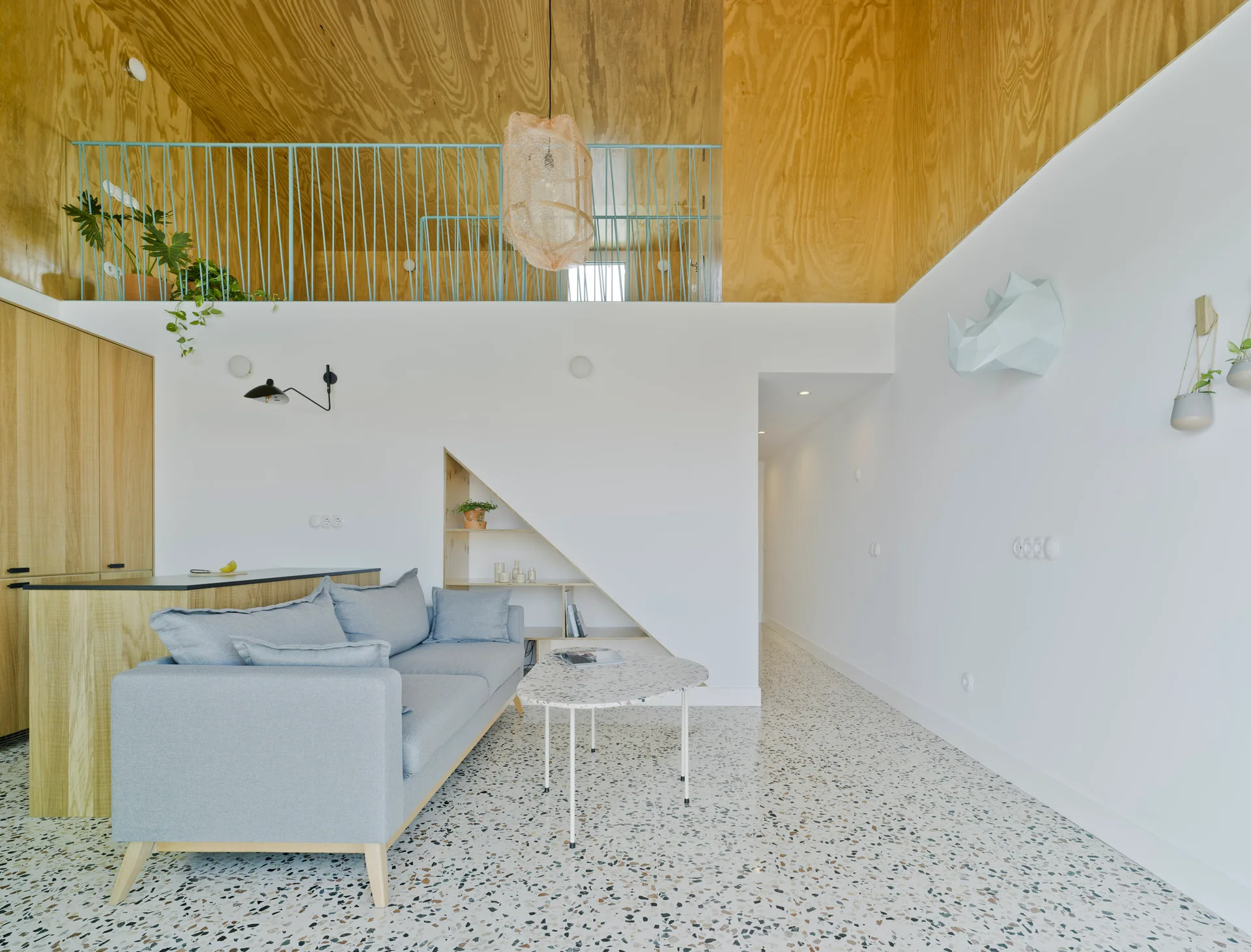 A minimalist living room featuring a grey sofa, speckled terrazzo flooring, and a plywood-clad mezzanine loft overhead.