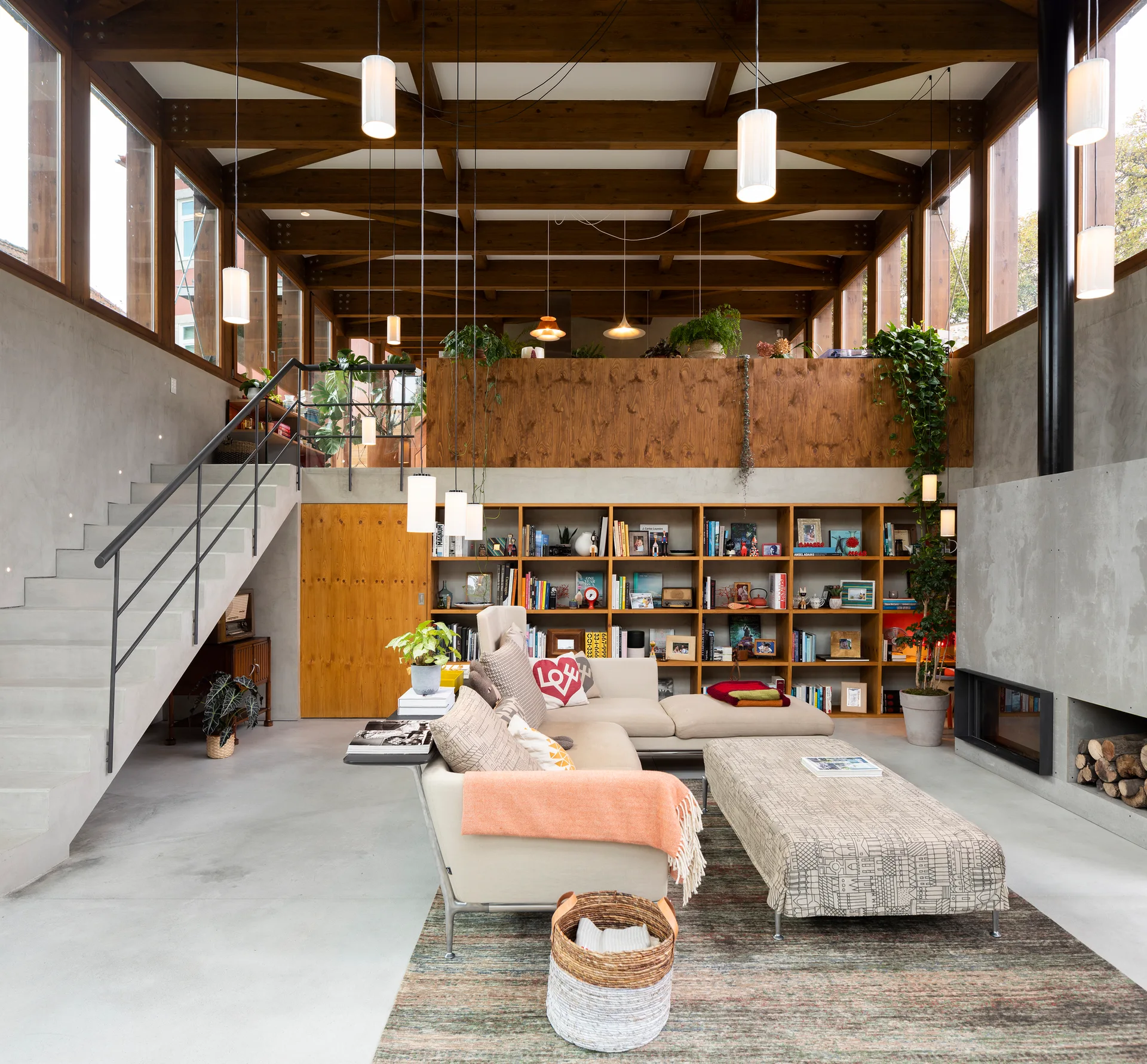 A modern double-height living room with concrete stairs, exposed timber ceiling beams, a built-in wooden bookcase, and a grey sectional sofa.
