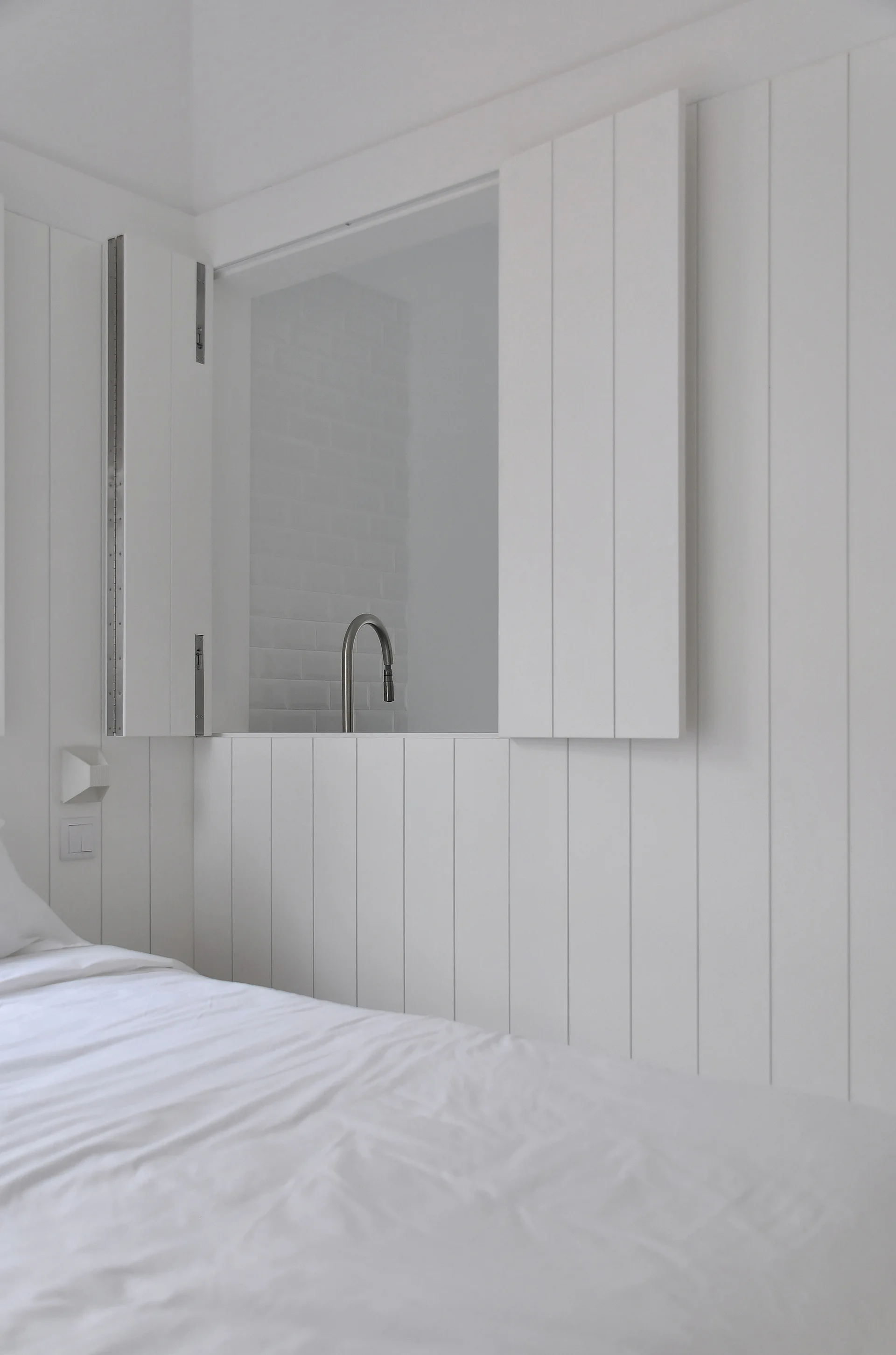 A minimalist white bedroom features vertical wall paneling and an open internal window with shutters showing a silver kitchen faucet.