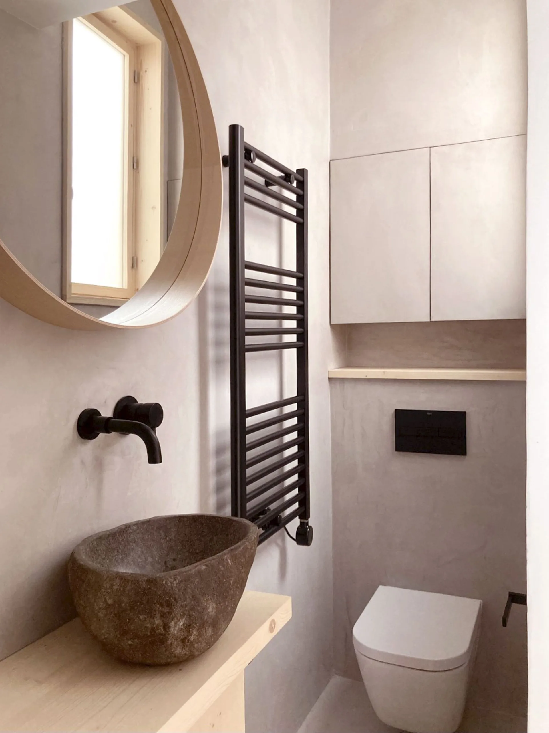 A minimalist powder room featuring a textured stone vessel sink on a wood shelf, matte black faucet, and a wall-hung toilet against grey plaster walls.