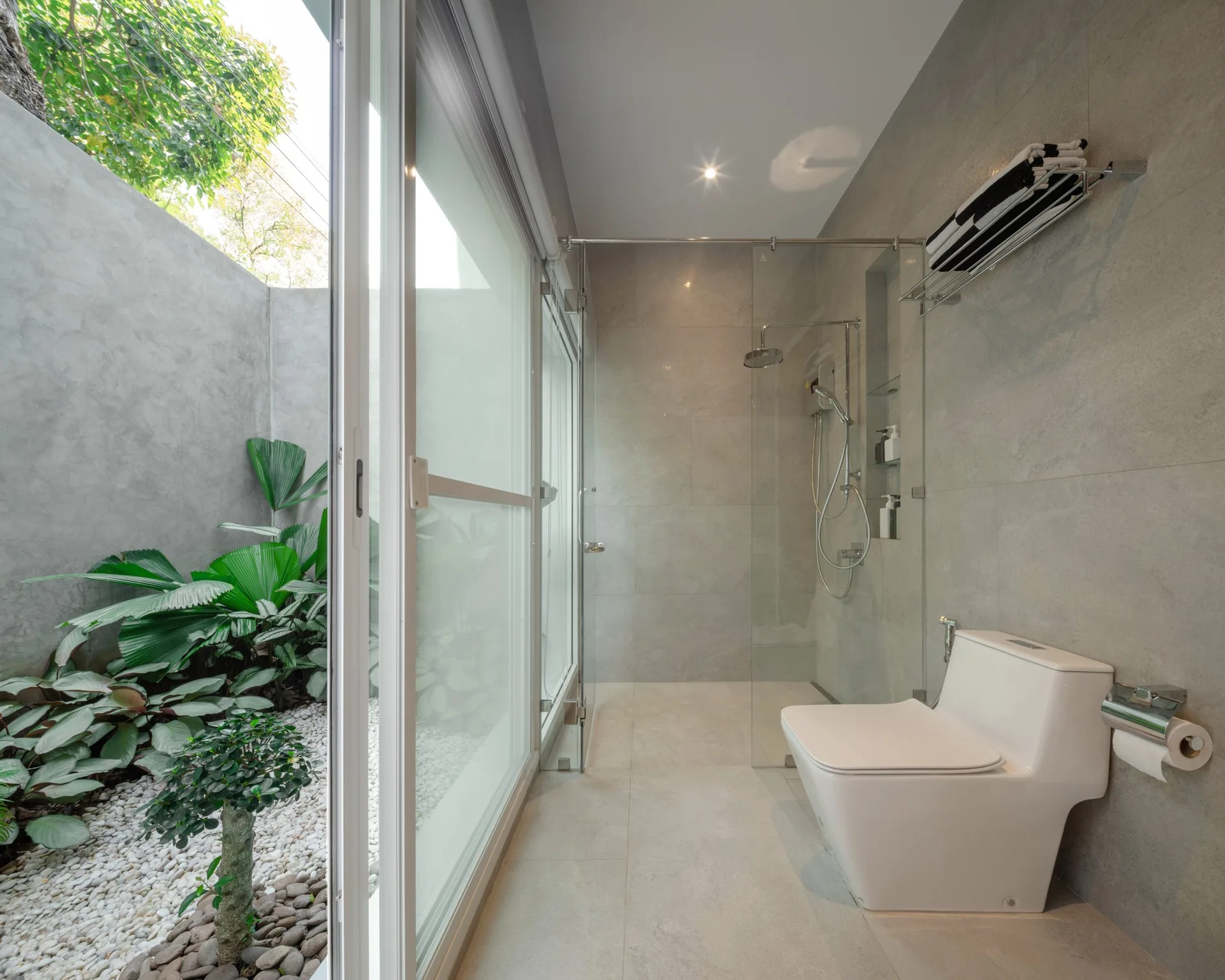 A modern gray tiled bathroom with a white toilet and glass shower door looking out to a small courtyard garden with pebbles and green plants.