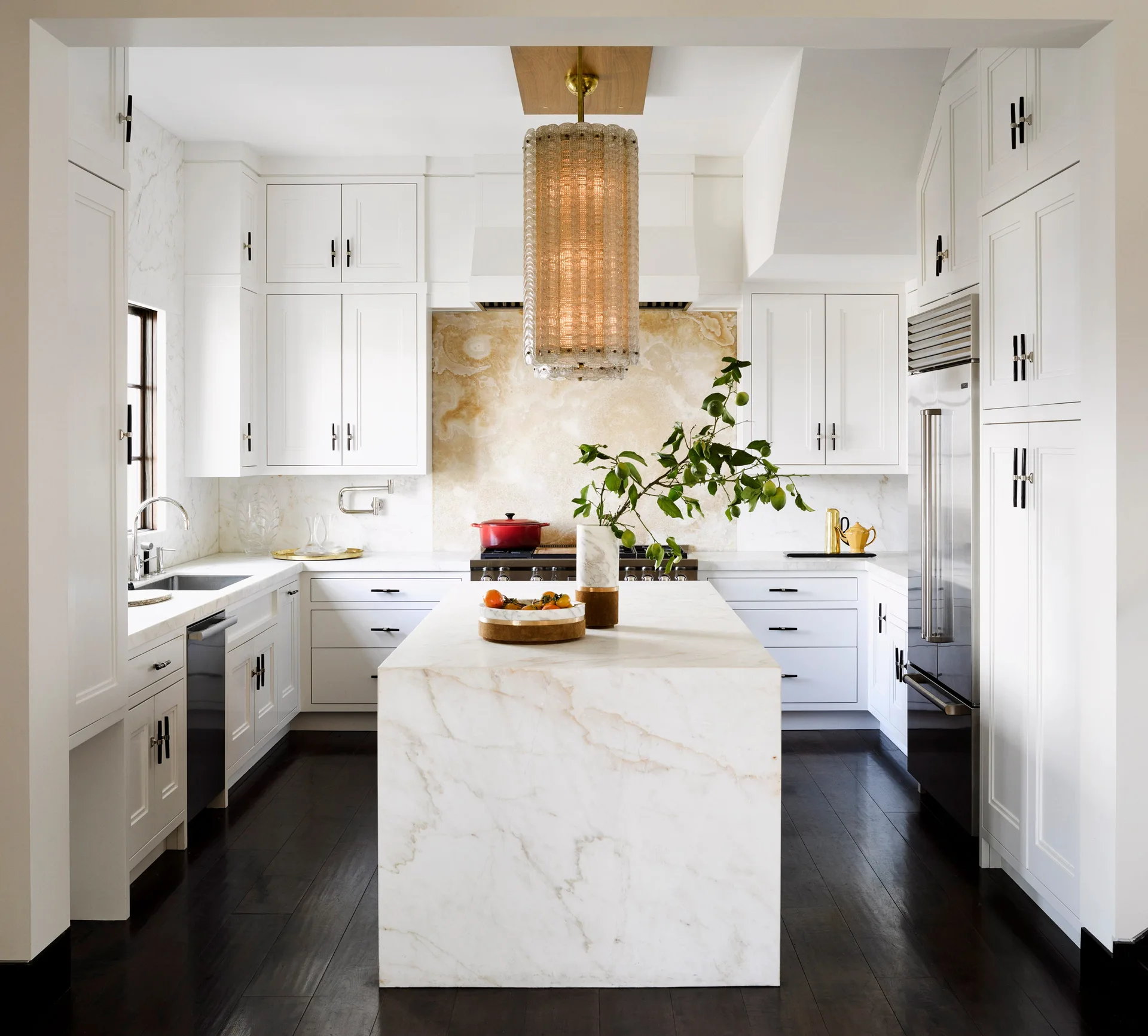 A symmetrical white kitchen features a central marble waterfall island, a gold stone slab backsplash, and a tall glass pendant light over dark floors.
