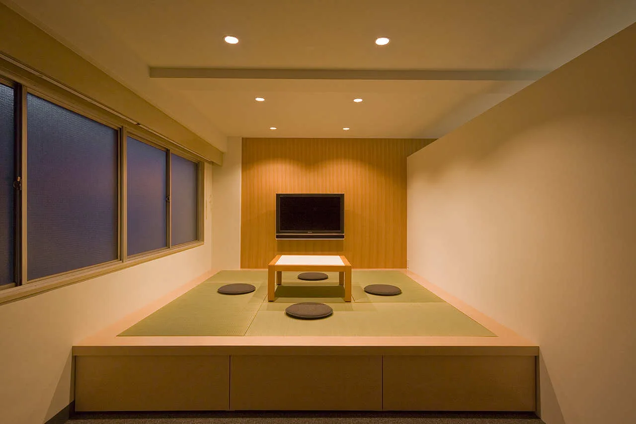 A minimalist living room with a raised wooden platform featuring green tatami mats, a low square table with grey cushions, and a wall-mounted TV.