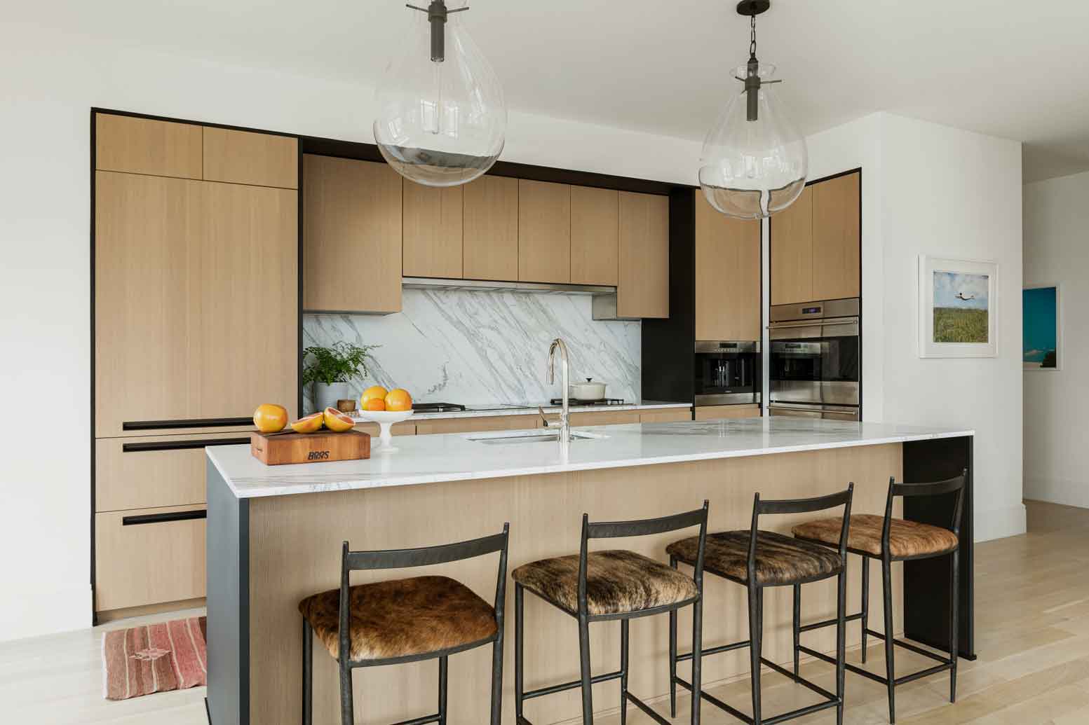 A modern kitchen features light wood cabinets framed in black, a marble slab backsplash, and glass globe pendant lights over an island with four stools.