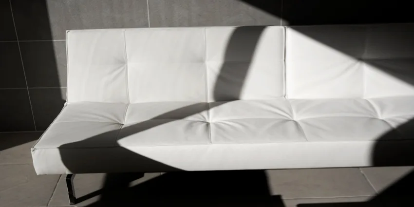 A minimalist white tufted sofa is positioned against a grey tiled wall with sharp geometric shadows across the seat.
