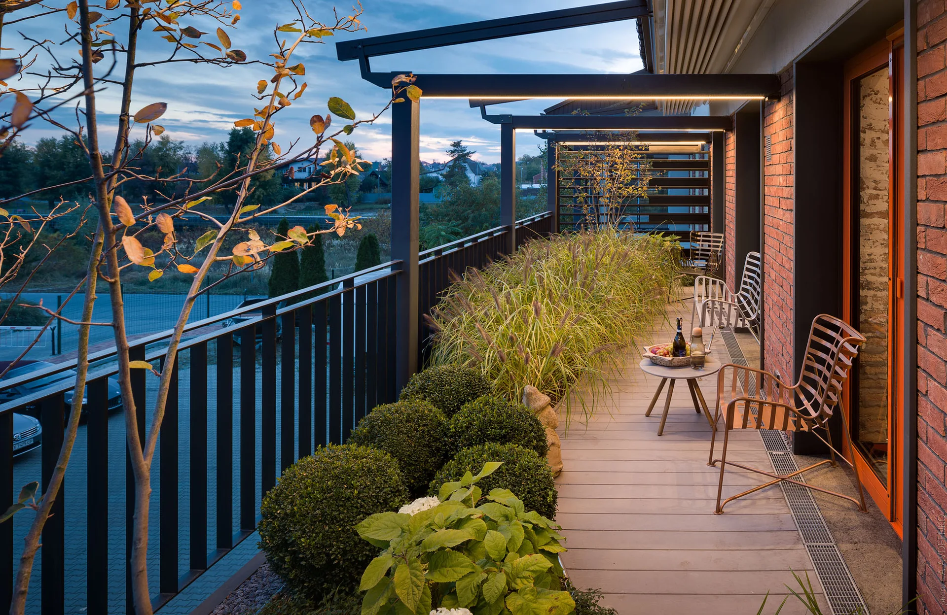 A long outdoor balcony features a black metal pergola with linear light strips, a brick wall, slat chairs, and a row of green plants.