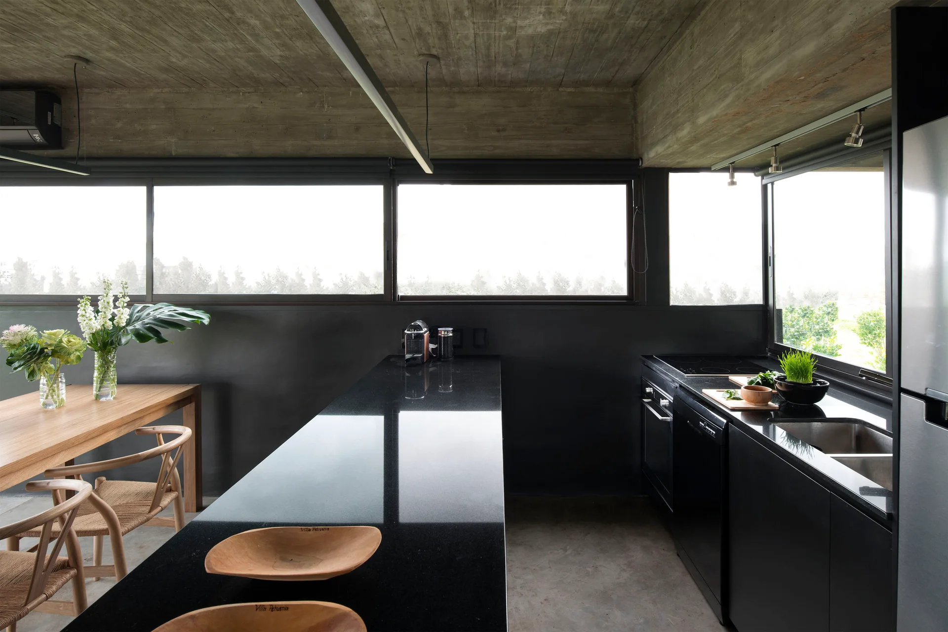 A modern kitchen features a polished black granite island, black cabinets, and a raw concrete ceiling alongside a wooden dining table and chairs.