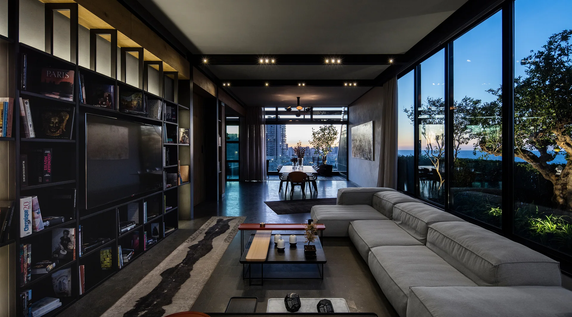 A modern open-plan living room with a grey sectional sofa, black metal bookshelves, and floor-to-ceiling windows overlooking a city at twilight.