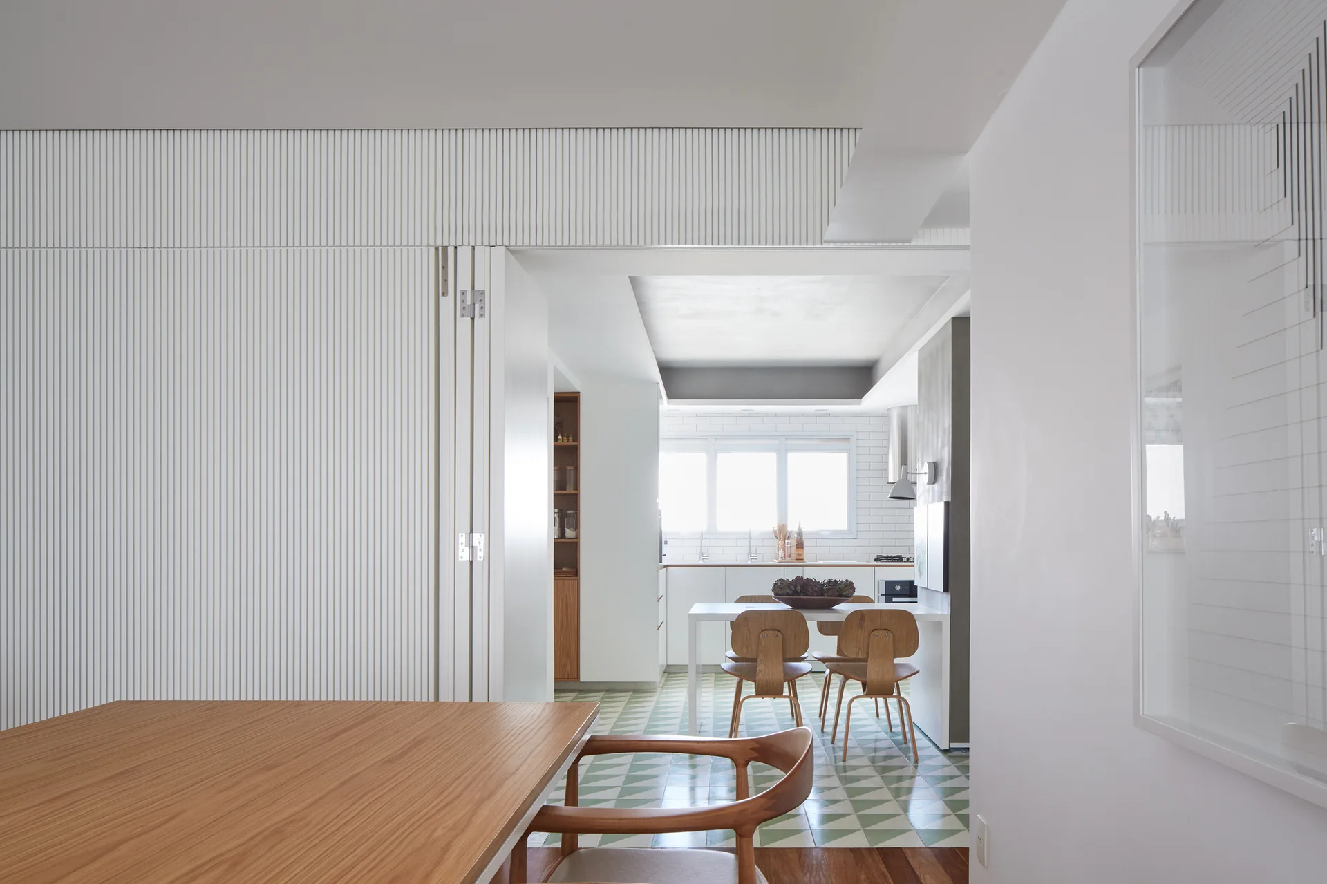 A dining area and kitchen featuring a white fluted wall partition, green and white geometric floor tiles, and mid-century wooden chairs.