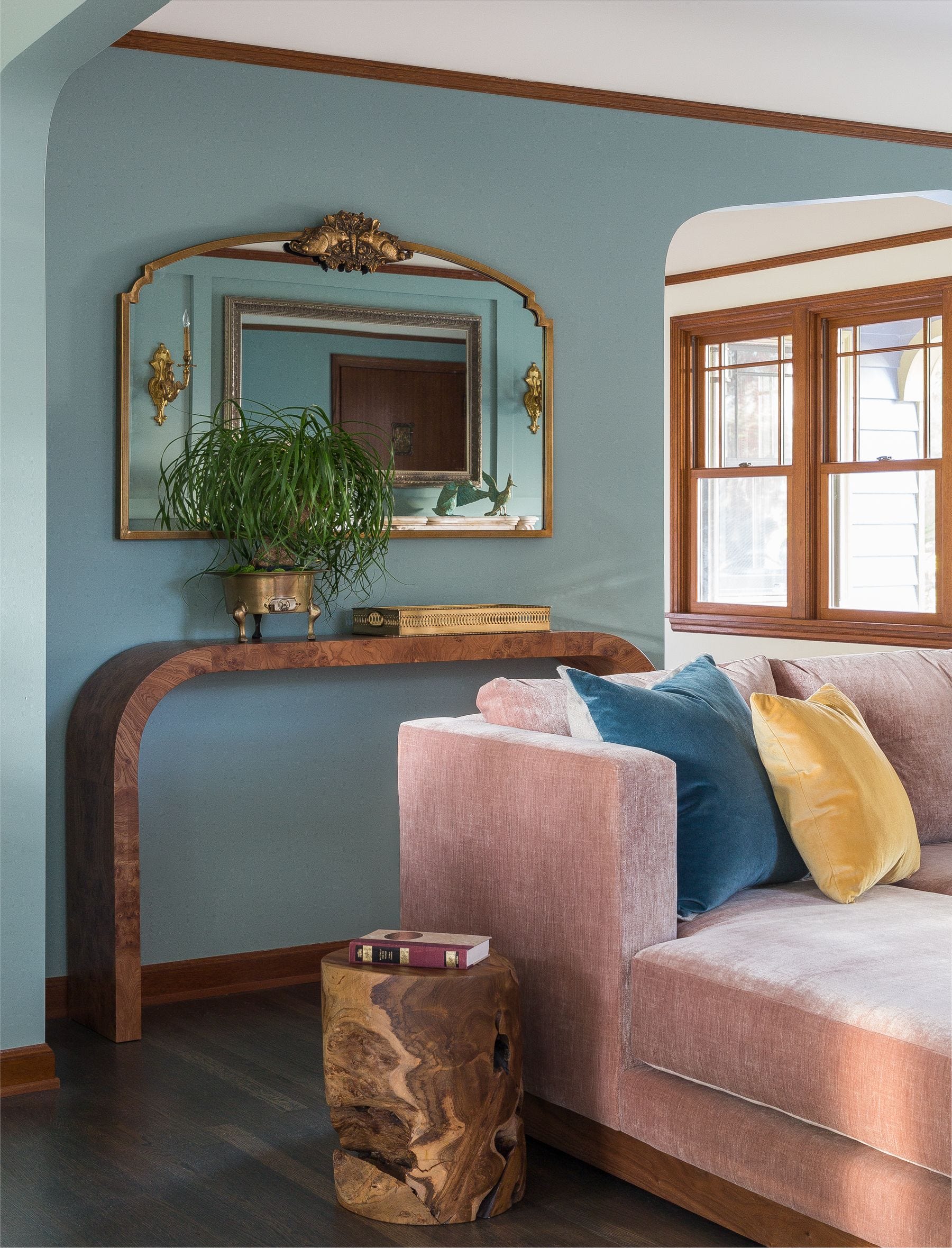 A living room with a pink velvet sofa, a curved burl wood console table against a muted blue wall, and an ornate gold mirror.