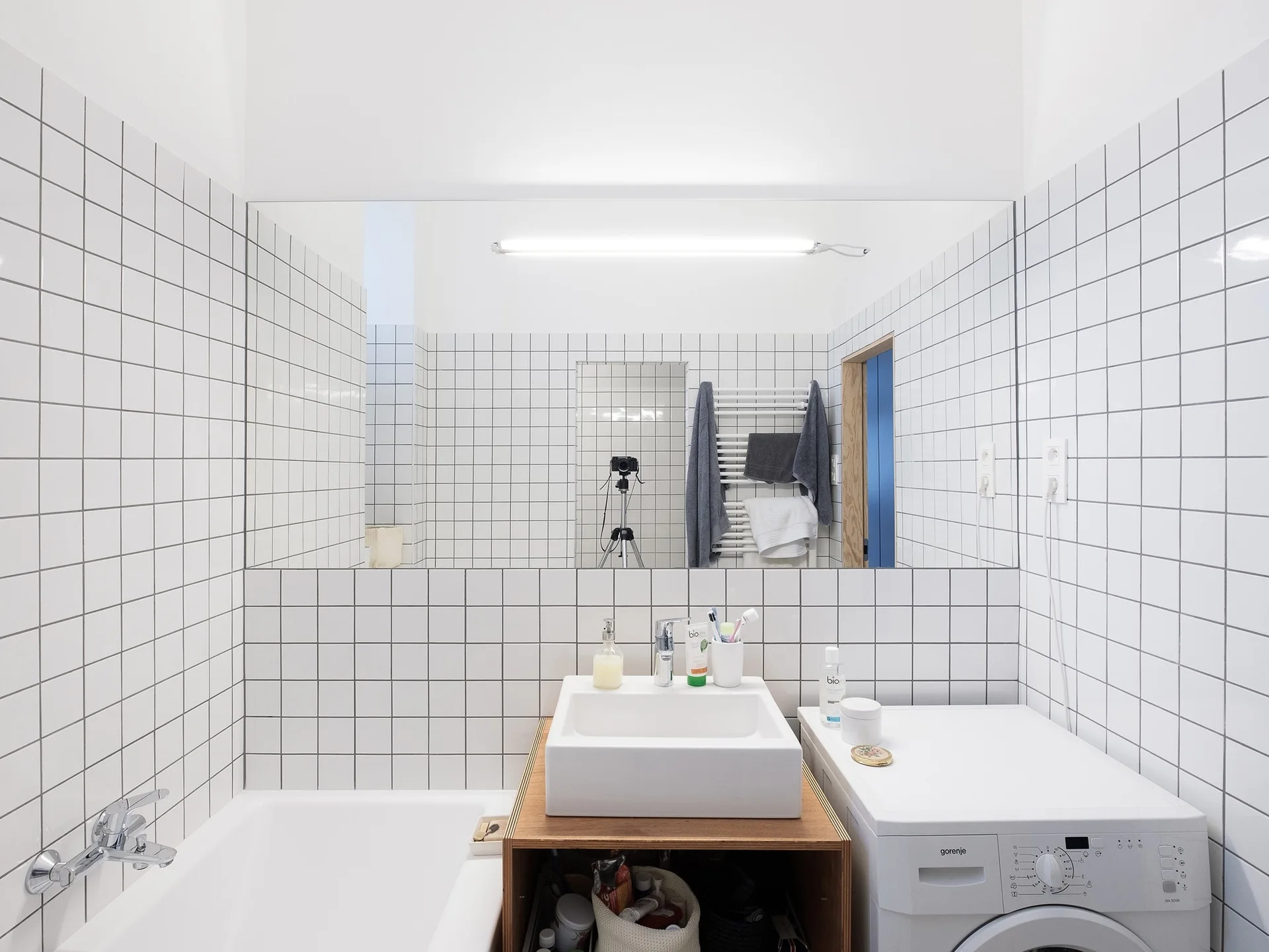 A minimalist bathroom with white grid wall tiles, a square washbasin on a wooden vanity, a large mirror, and an integrated washing machine.