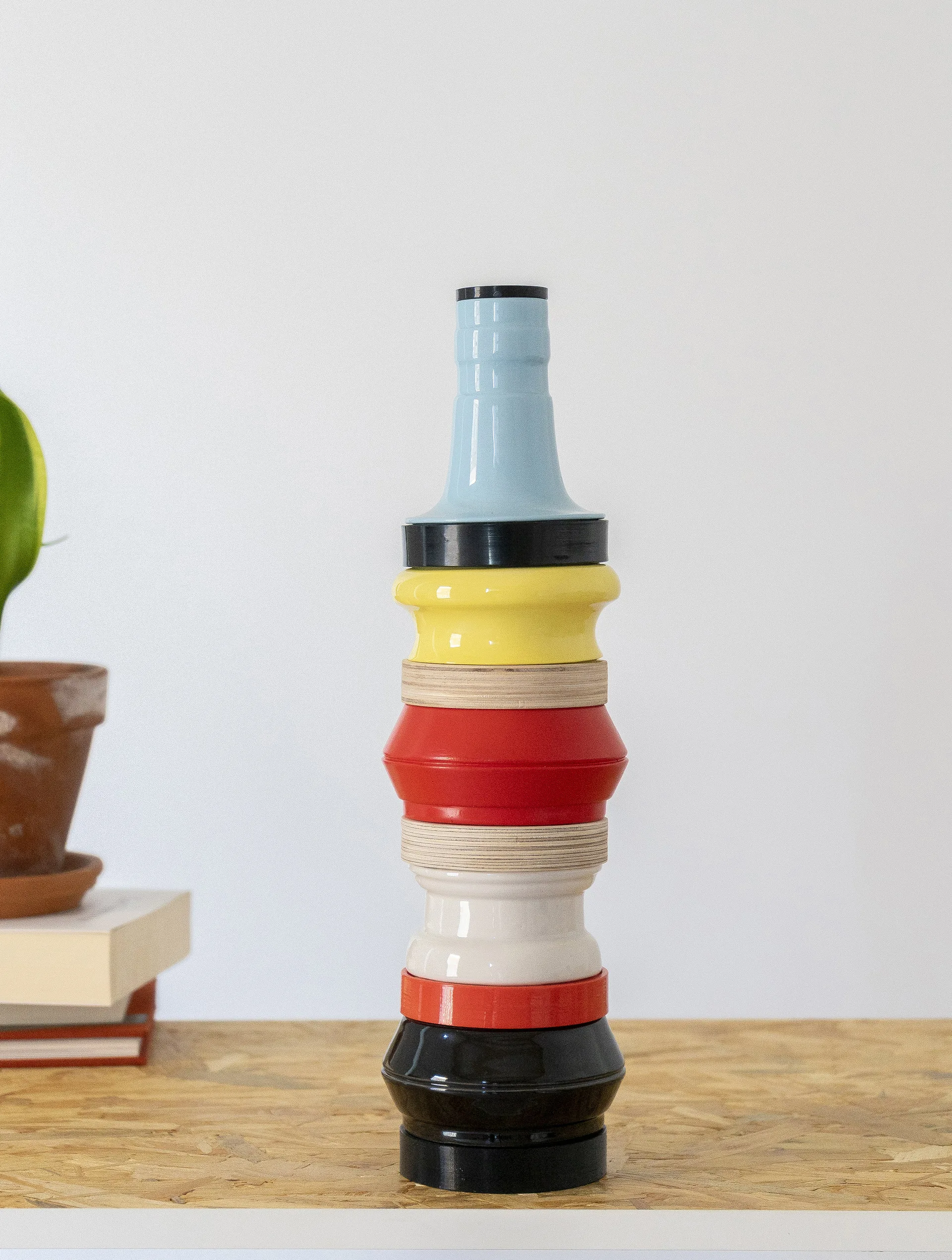 A multicolored stacked geometric ceramic sculpture sits on a raw wood tabletop next to a stack of books and a small potted plant.