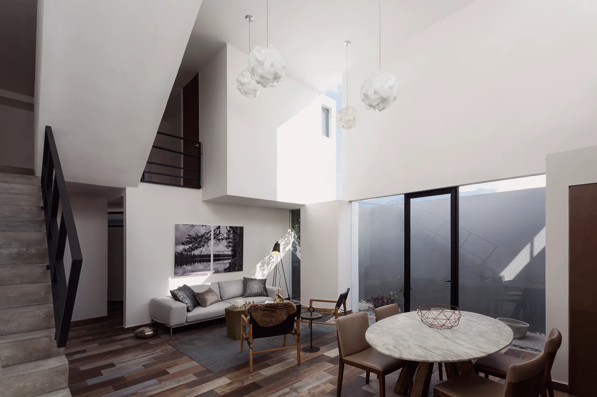 A bright double-height living room features a light grey sofa, concrete stairs, and a round marble dining table beneath sculptural white pendant lights.