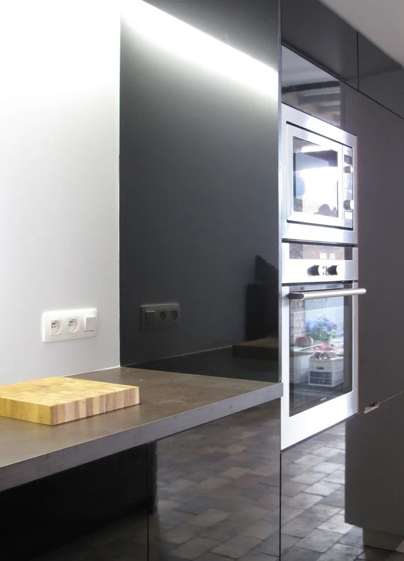 A modern kitchen corner with reflective high-gloss black cabinets, a built-in stainless steel oven stack, and a wooden cutting board.