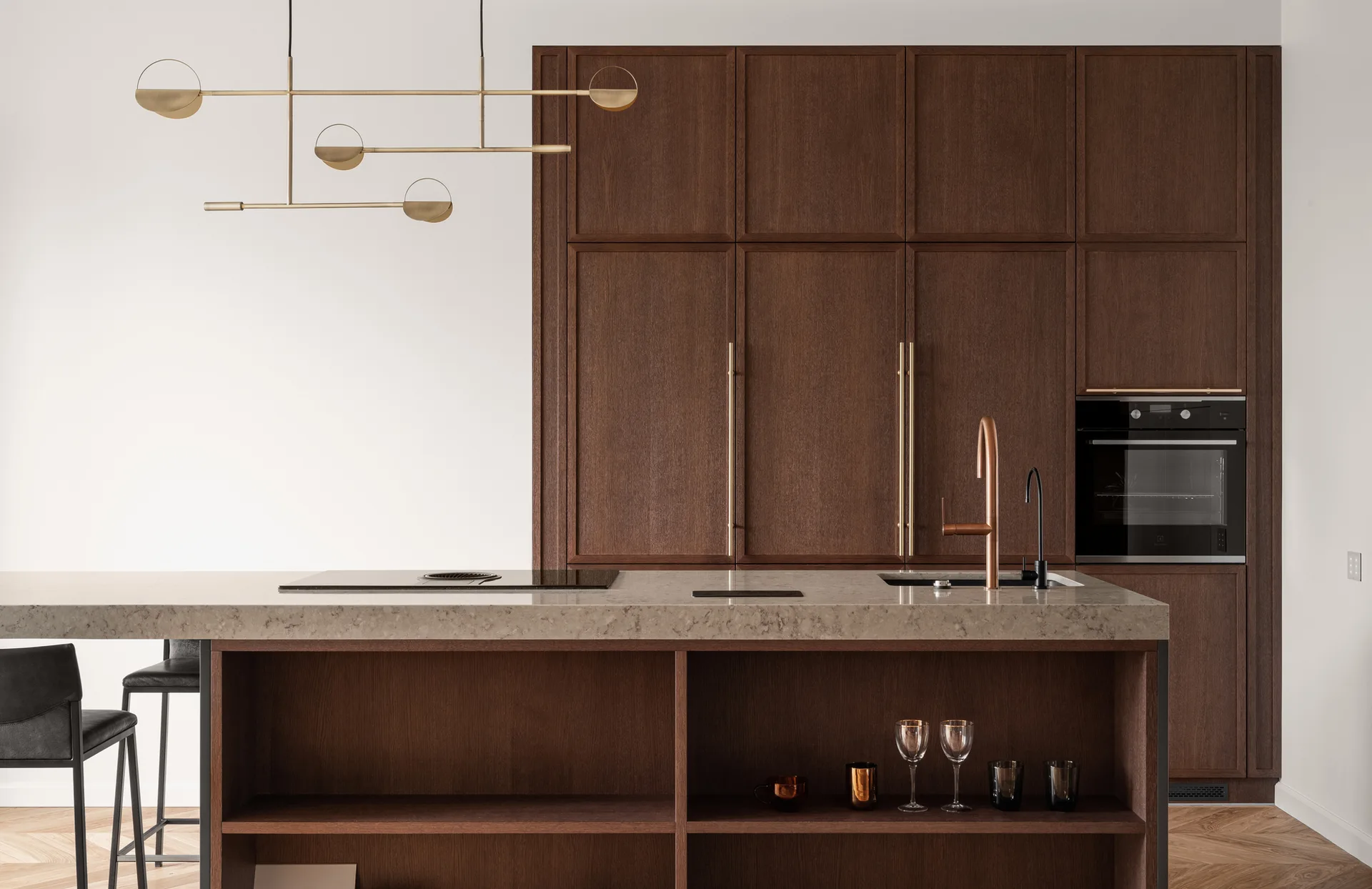 A minimalist kitchen featuring dark wood cabinetry, a stone-topped island with open shelving, and a modern gold linear pendant light.