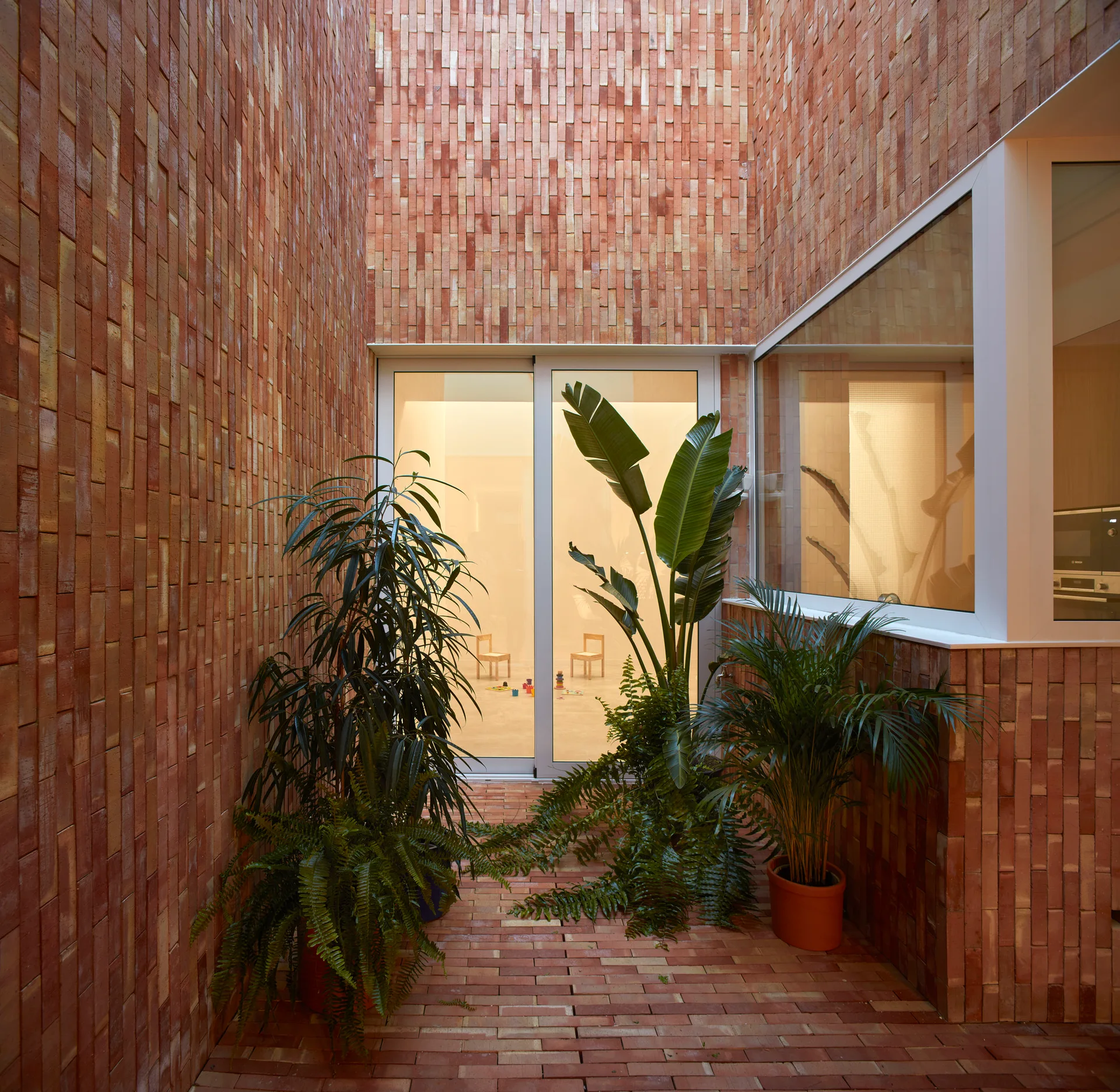 Tall terracotta brick walls enclose a small courtyard patio paved in matching brick, featuring several large potted green plants and glass sliding doors.