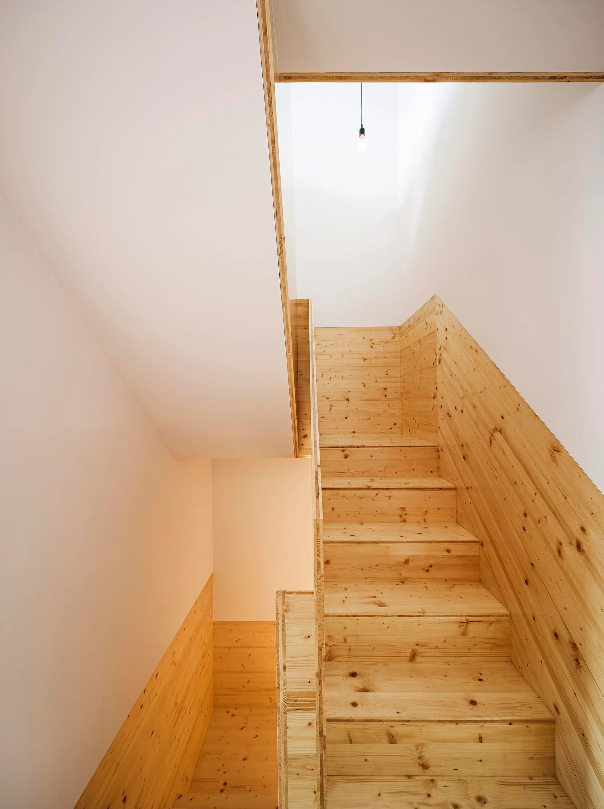 A minimalist wooden staircase built with knotty pine rises between white walls under natural light from a skylight above.