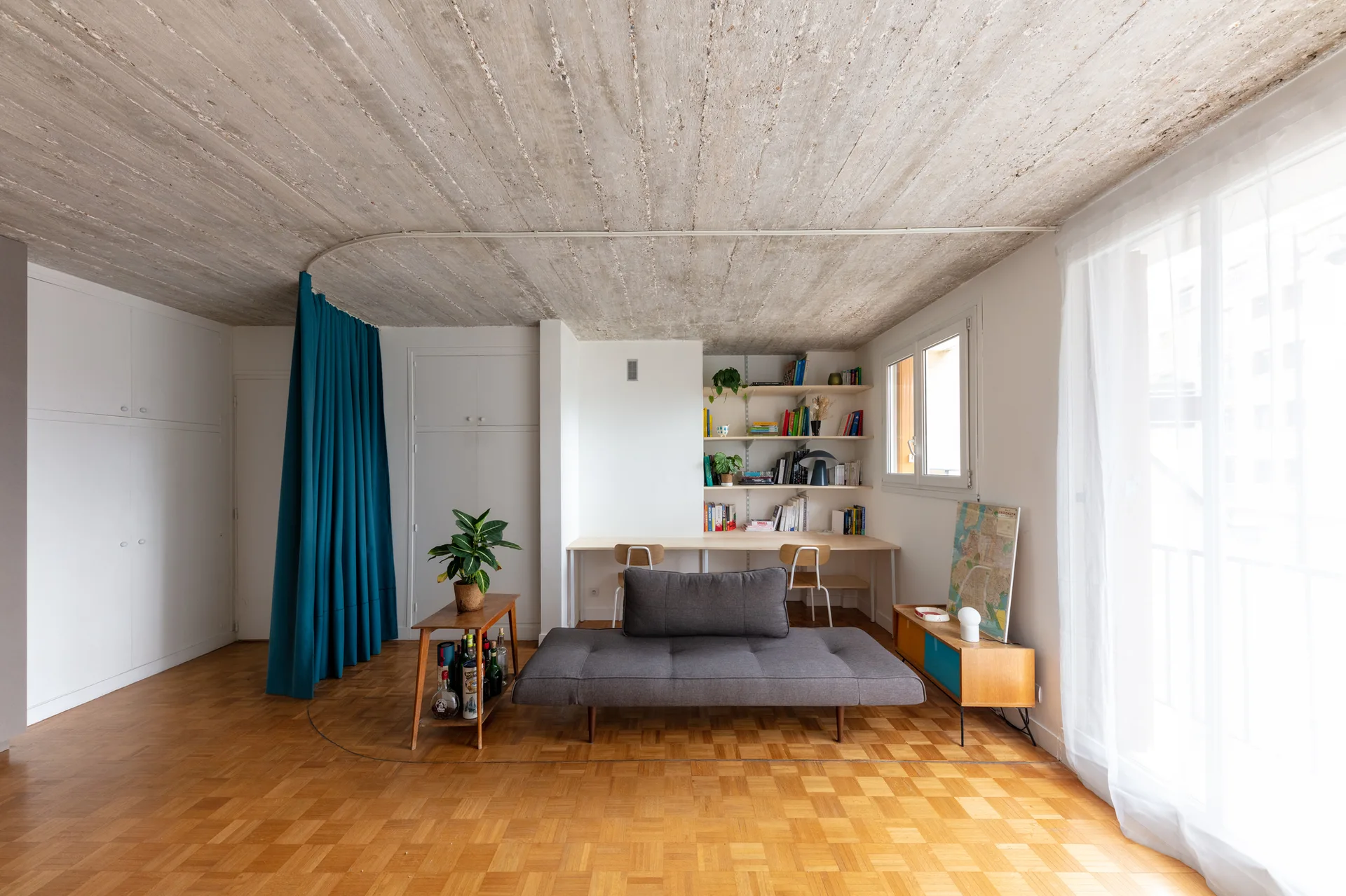 A grey sofa bed sits in a living room featuring an exposed concrete ceiling, parquet wood flooring, and a floor-to-ceiling teal room divider curtain.