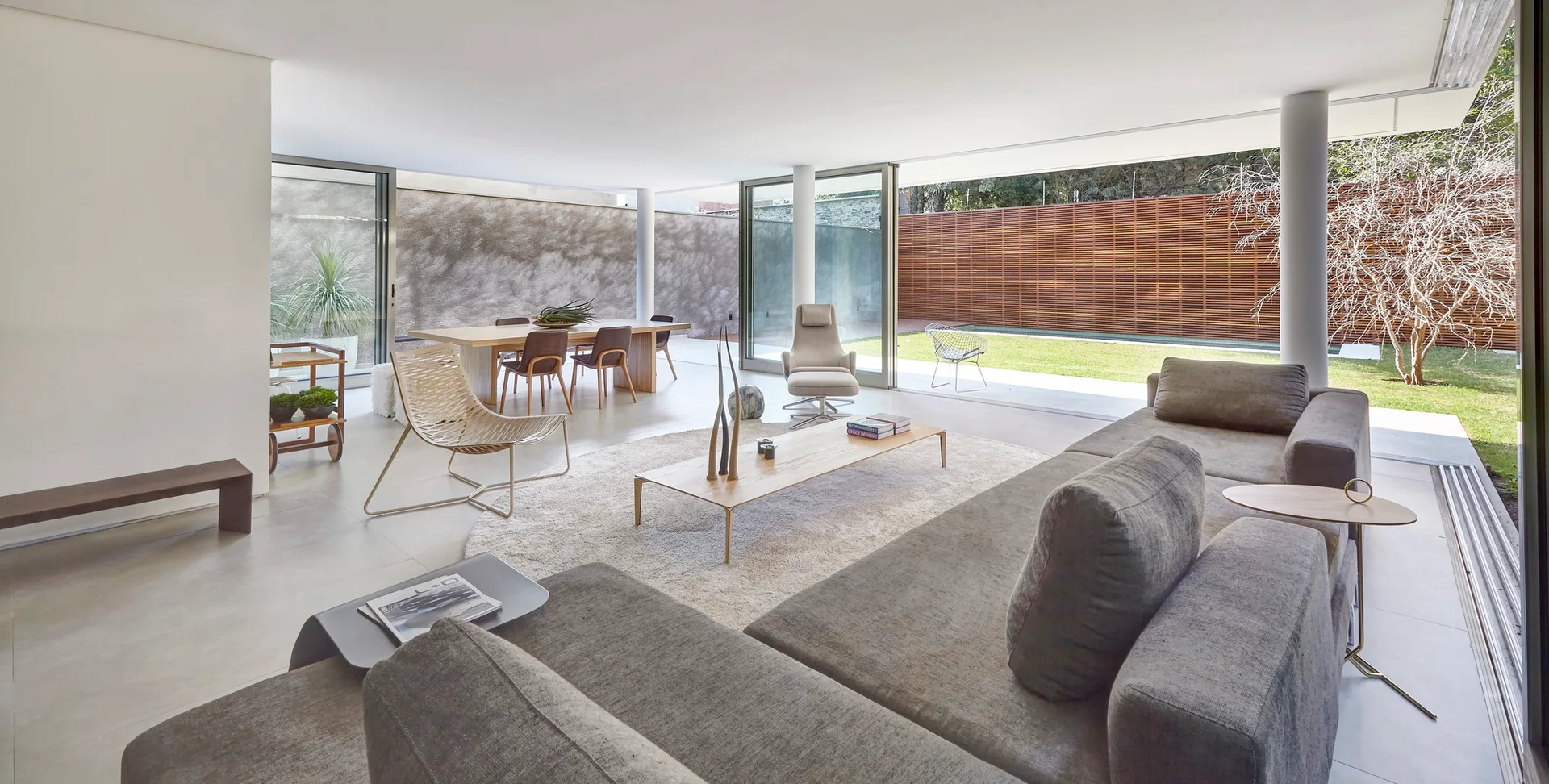 A minimalist open-concept living room features a grey sectional sofa, low wooden coffee table, and sliding glass doors opening to a grassy yard.