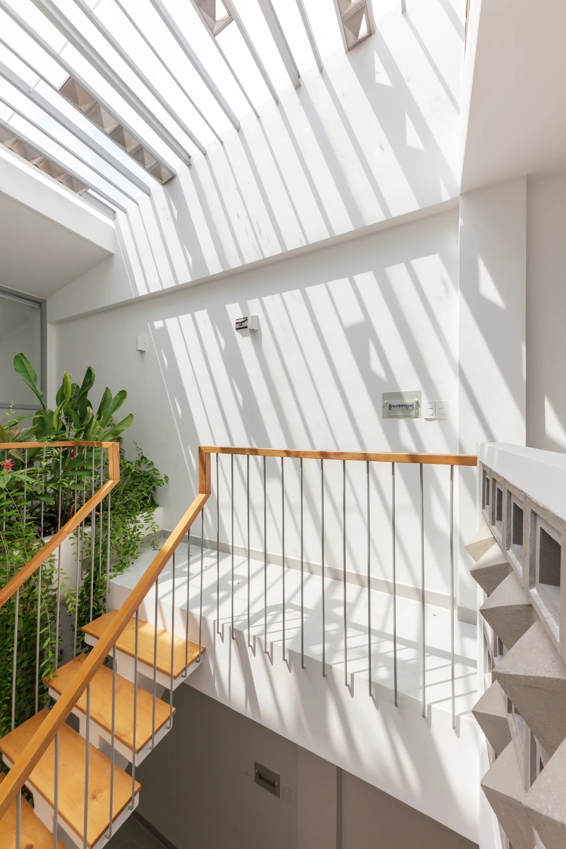A modern wooden staircase with a metal railing sits under a slatted ceiling that projects linear striped shadows onto a white wall.