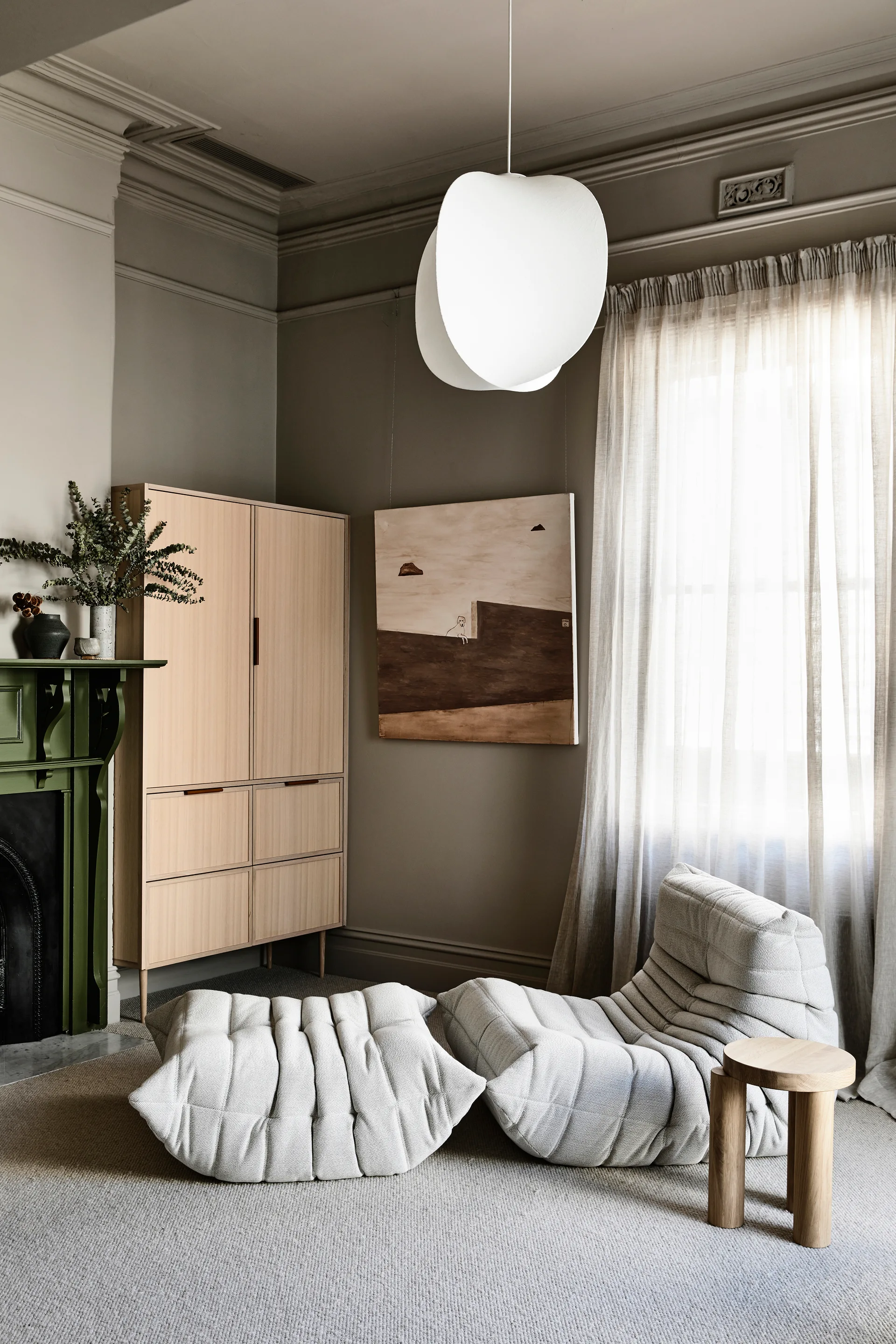 A low-profile tufted gray armchair and matching ottoman sit in a neutral room near an olive green mantelpiece and light wood cabinet.