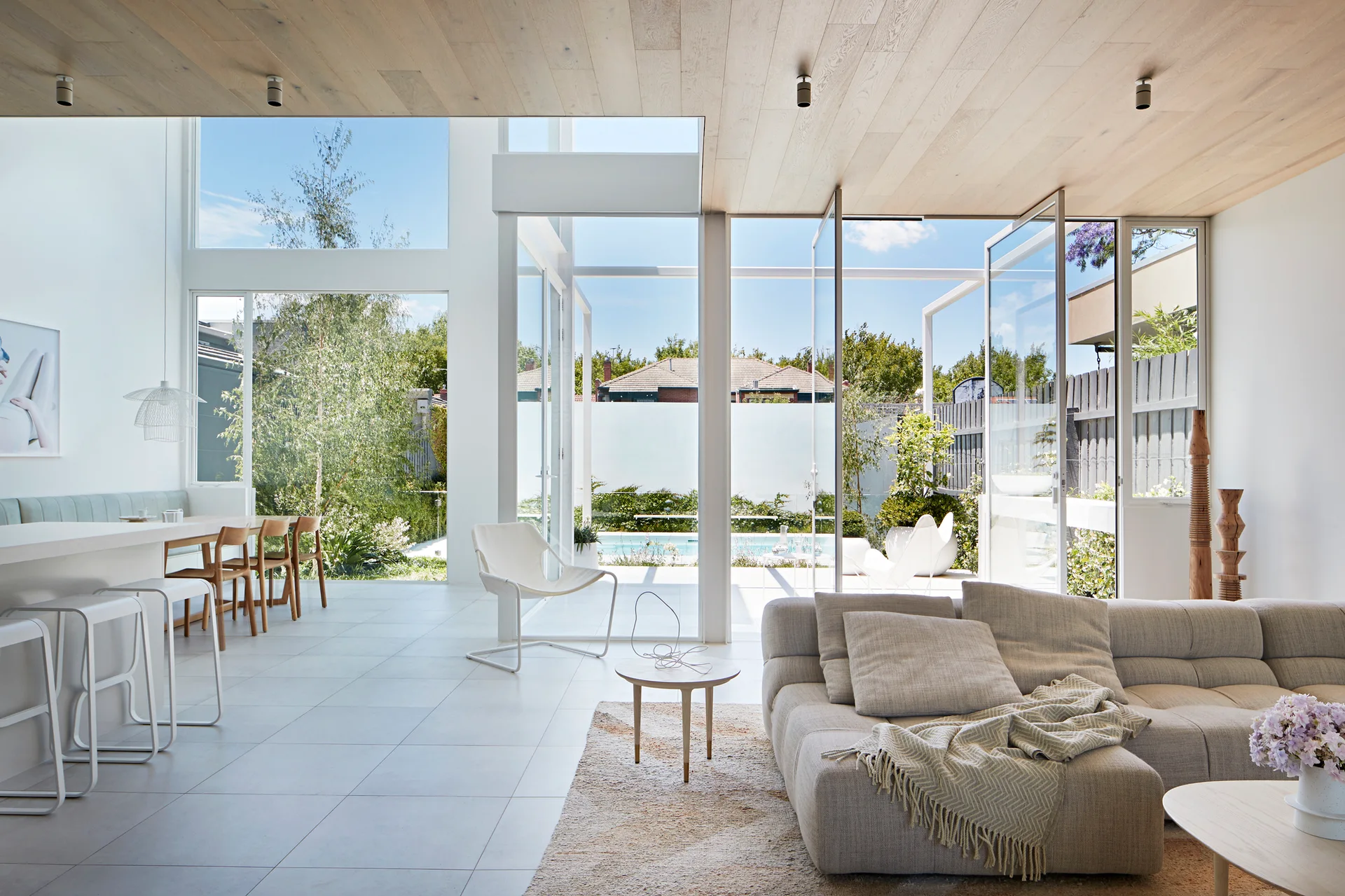 A modern living room features a beige modular sofa, white tile floors, a light wood paneled ceiling, and large glass doors opening to a backyard pool.