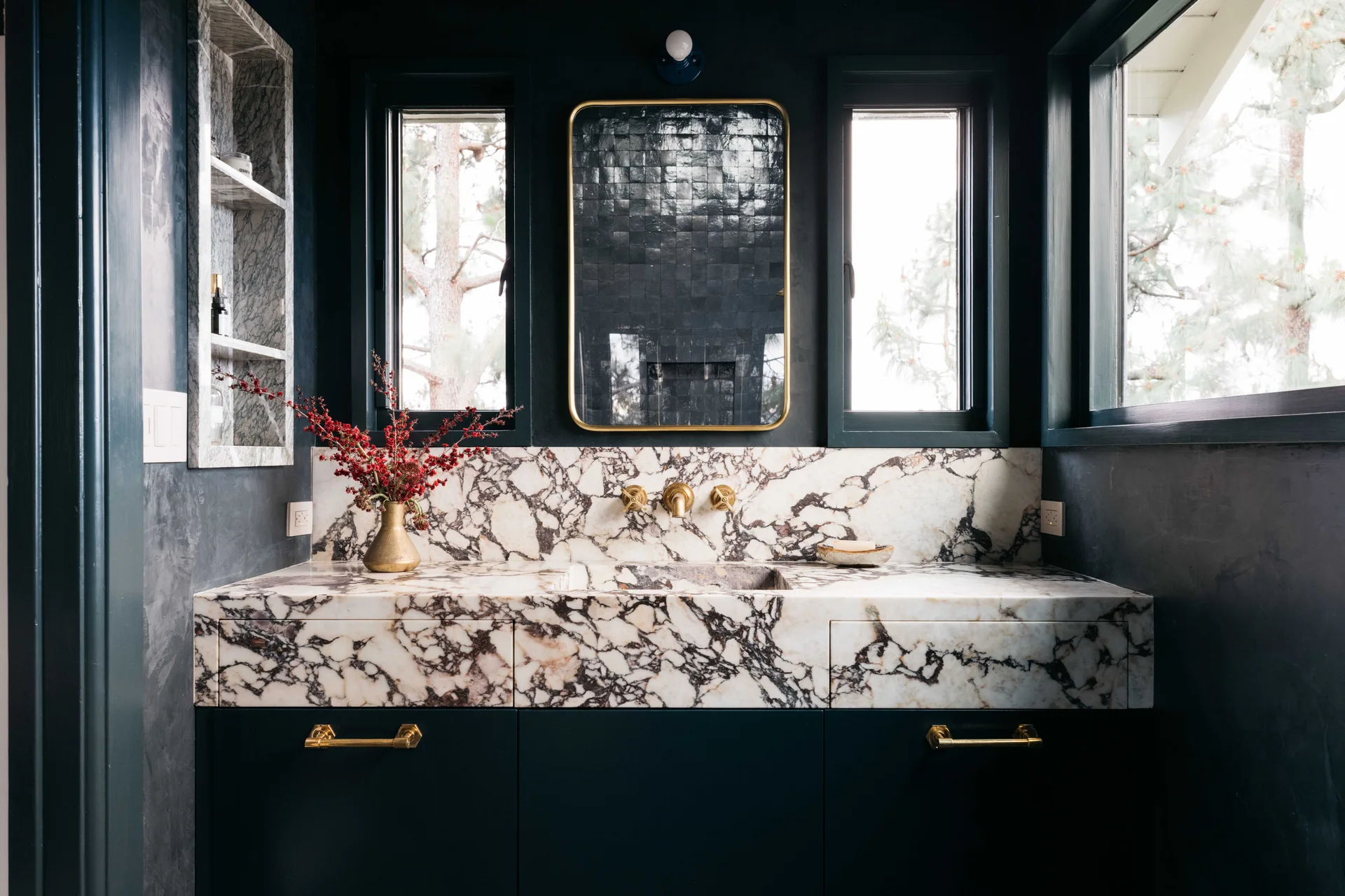 A dark navy bathroom features a heavily veined marble vanity with an integrated sink, brass hardware, and a brass-framed mirror flanked by two windows.